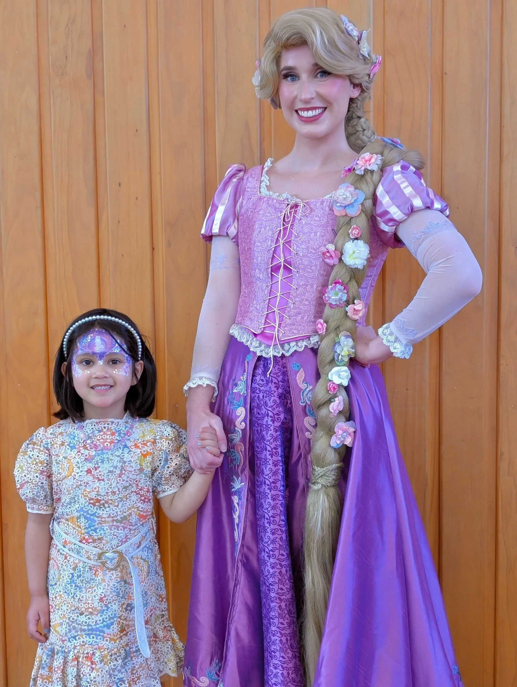 A woman dressed as Rapunzel from Disney's Tangled, holding hands with a young girl with face paint and a dress, standing in front of a wooden wall.