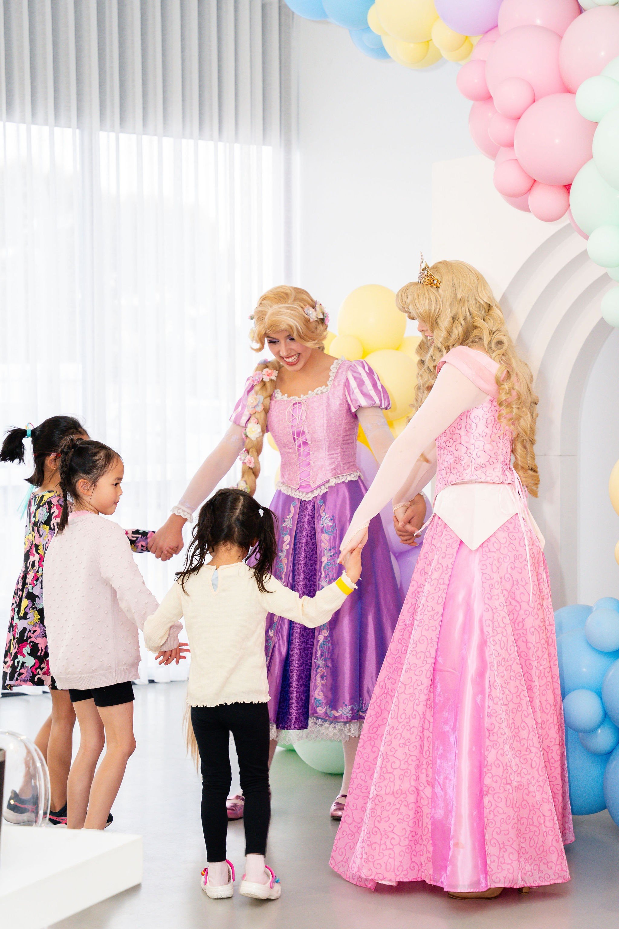 Children celebrating with two women dressed as princesses, holding hands and playing together, surrounded by pastel-colored balloons.