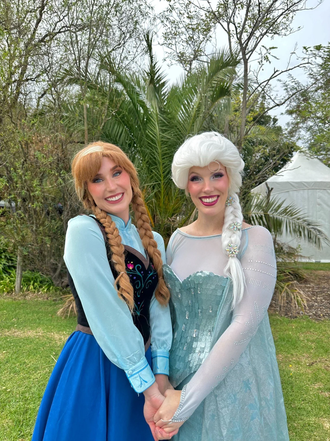 Two women dressed as Disney characters Elsa and Anna from Frozen, standing outdoors on grass with trees and plants in the background, holding hands and smiling.