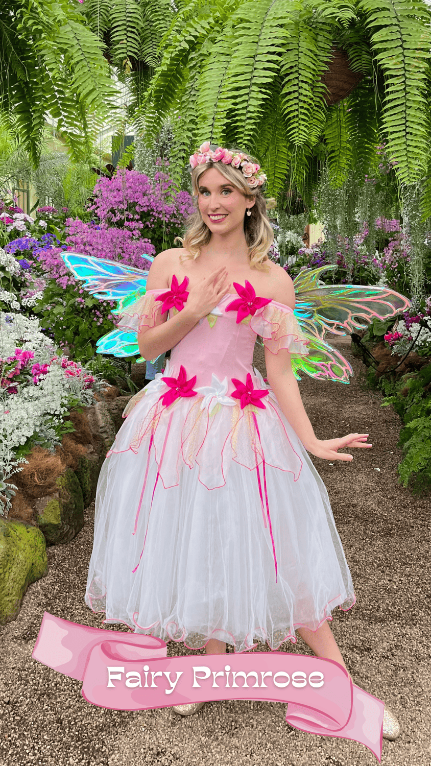 A young woman dressed as a fairy princess in a pink and white dress with large pink flowers and iridescent fairy wings, standing in a lush garden with various colorful flowers and green foliage. She has blonde hair adorned with a pink floral crown, a