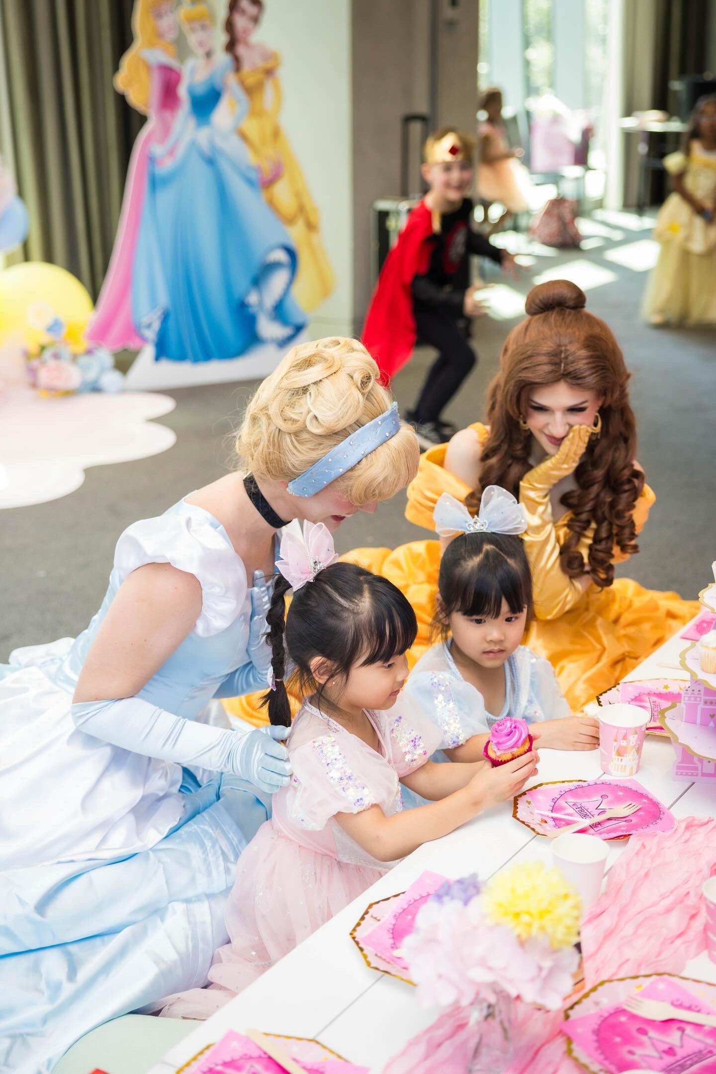 Children at a princess-themed birthday party, with life-sized character cutouts and princess decorations, celebrating with cupcakes.