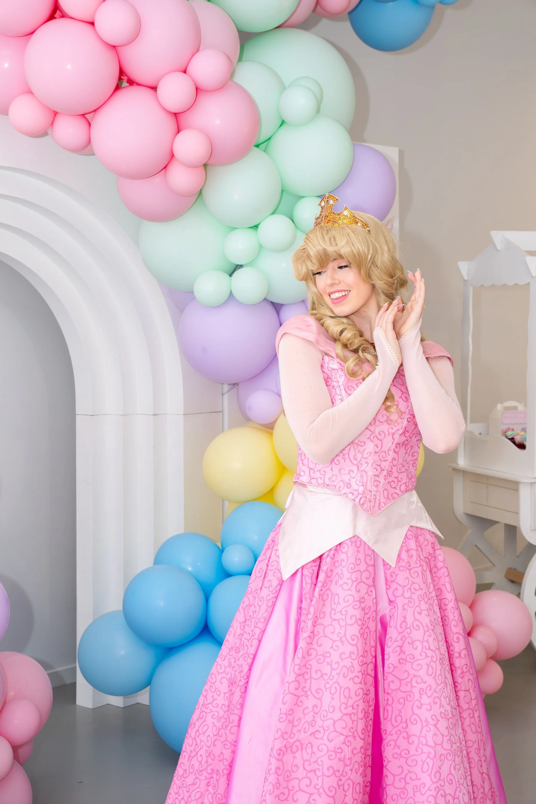 A woman dressed as a princess in a pink gown and wearing a crown, standing in front of colorful balloons arranged in a decorative arch.