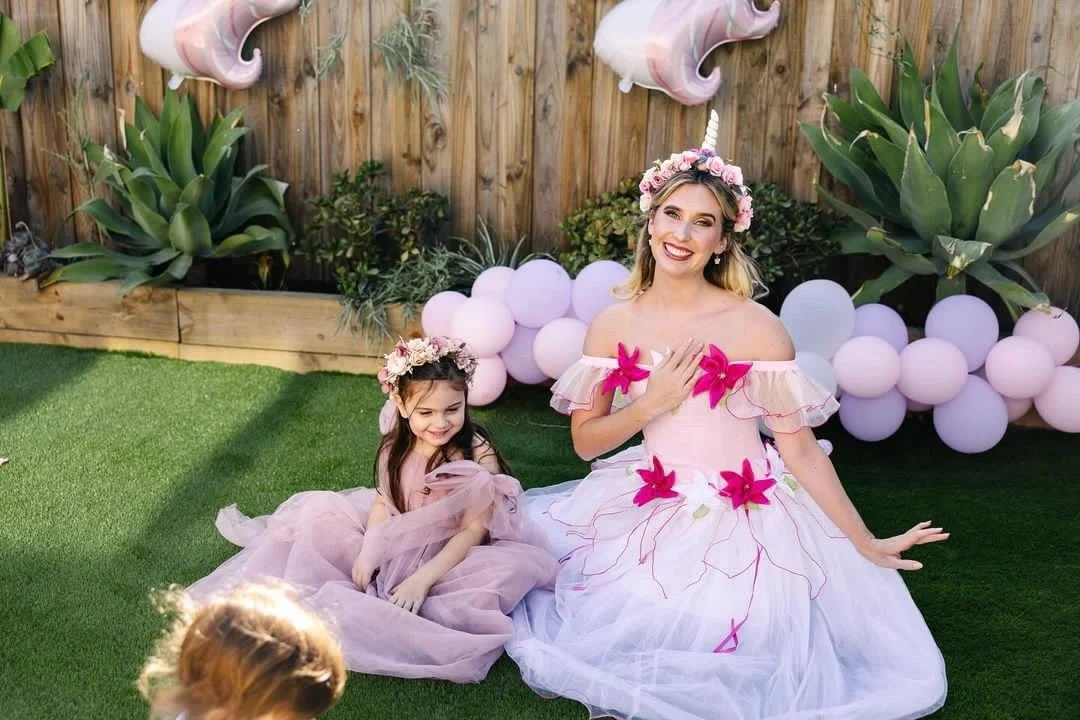 Woman and young girl dressed in pink fairy costumes, sitting on green grass at a birthday party with pink balloons and plants in the background.