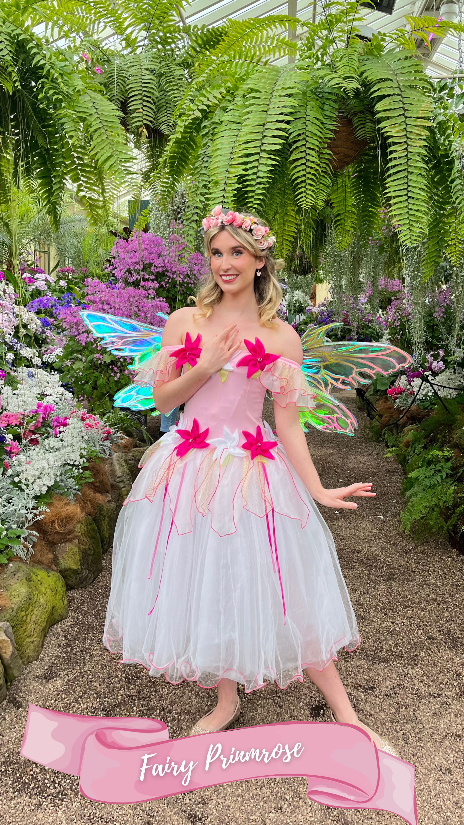 A young woman dressed as a fairy princess in a pink and white dress with large pink flowers and iridescent fairy wings, standing in a lush garden with various colorful flowers and green foliage. She has blonde hair adorned with a pink floral crown, a
