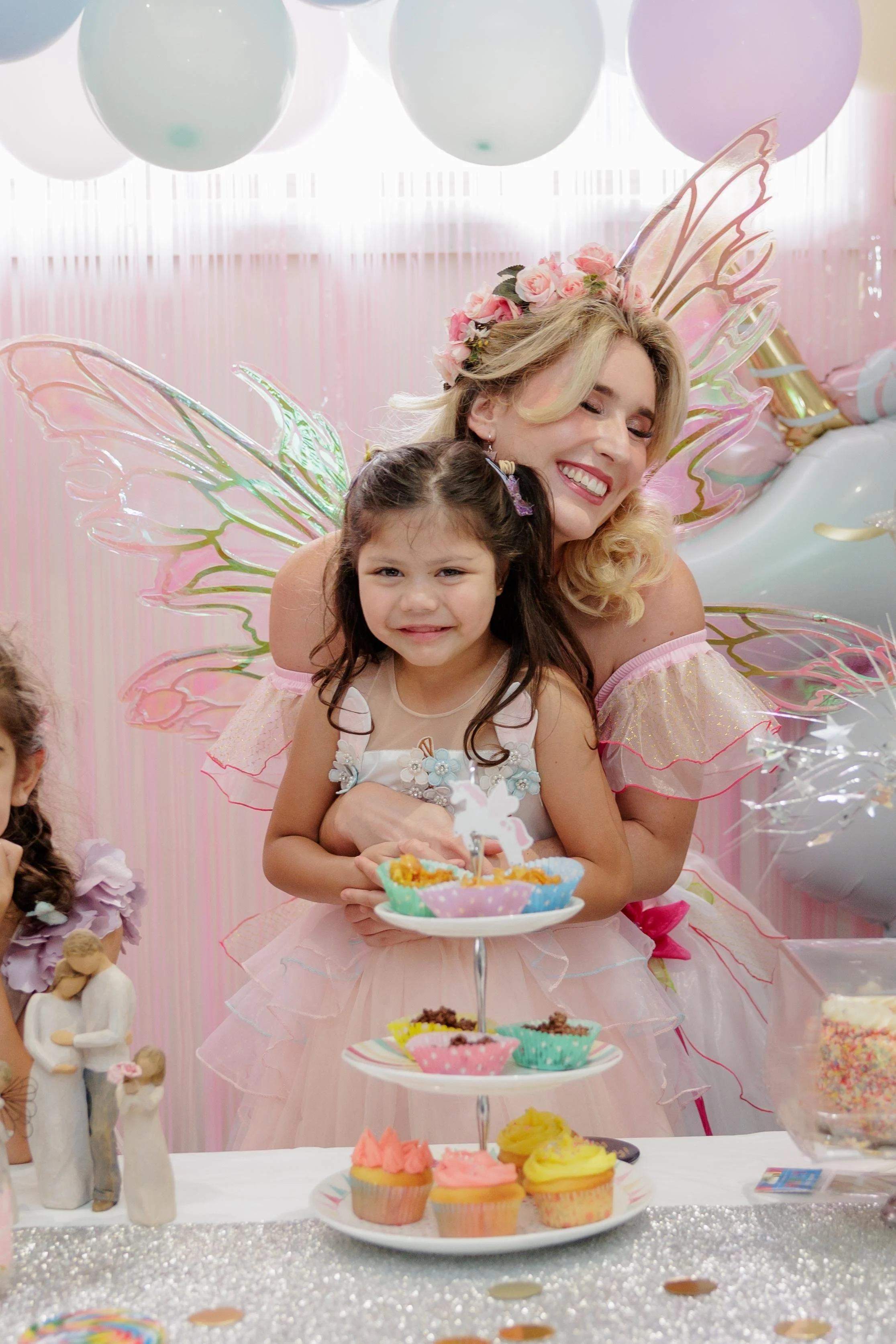 A woman dressed as a fairy with blonde hair, pink fairy wings, and a floral crown, hugging a young girl with long dark hair, wearing a fairy princess dress at a birthday party. The girl is smiling and holding a small cake, with a pink and purple themed birthday setup and cupcakes on the table.