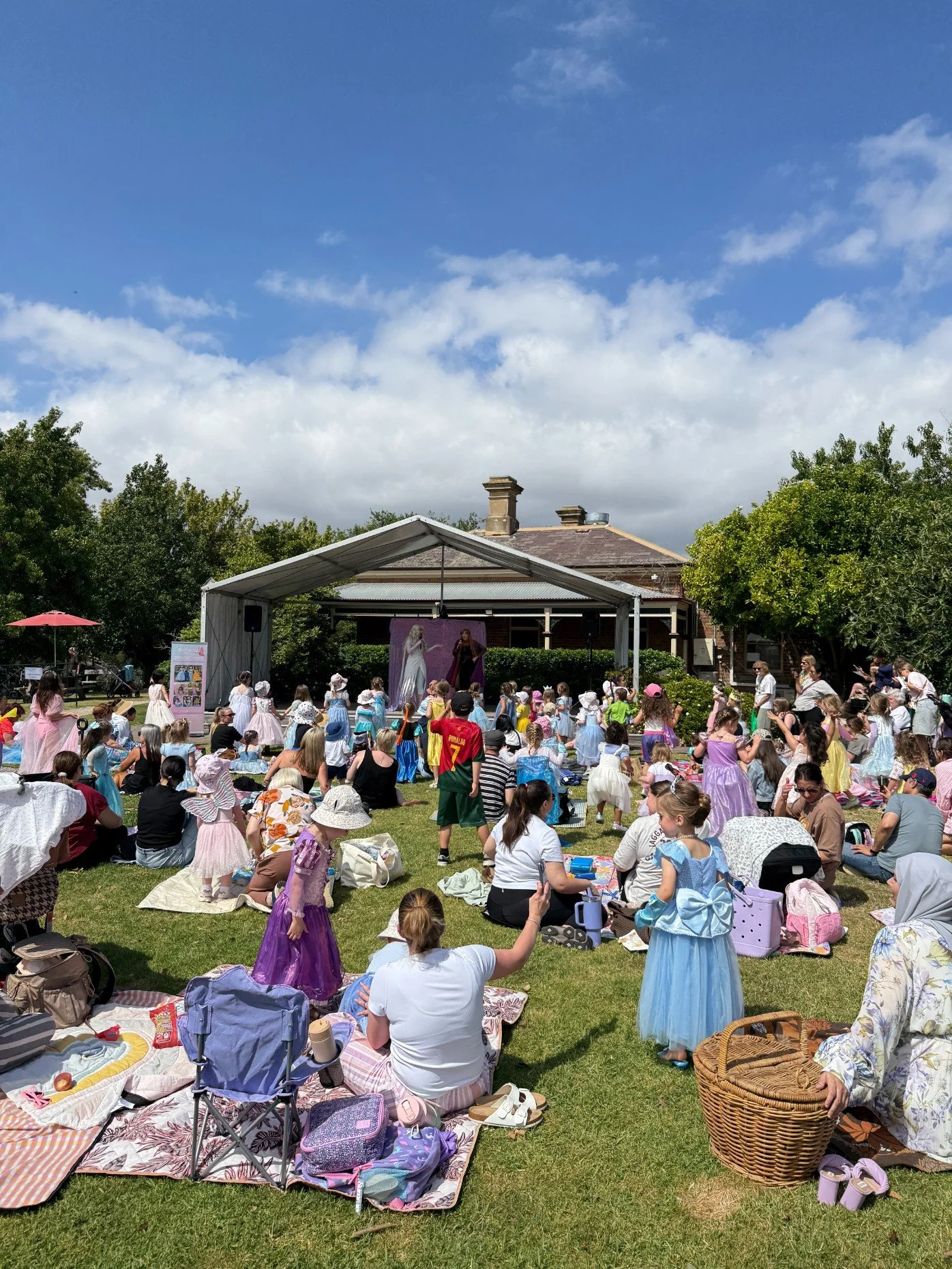 A large group of people, including many children, gathered outdoors on a grassy area for a live performance with a stage under a tent, with some children dressed in princess costumes, on a mostly sunny day with blue skies and a few clouds.