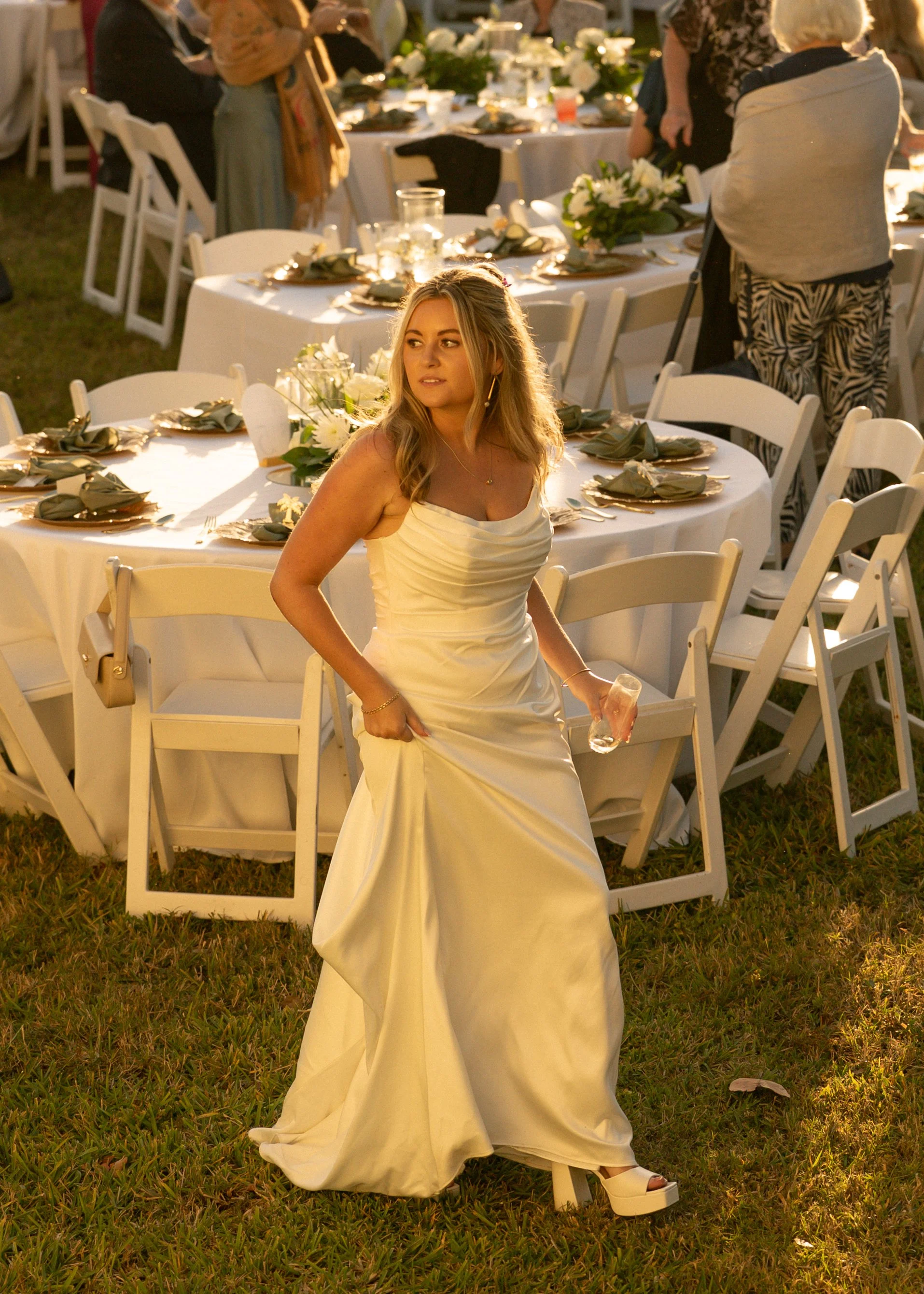 A young woman in a white wedding dress holding a drink is standing next to a decorated banquet table at an outdoor event, likely a wedding reception.