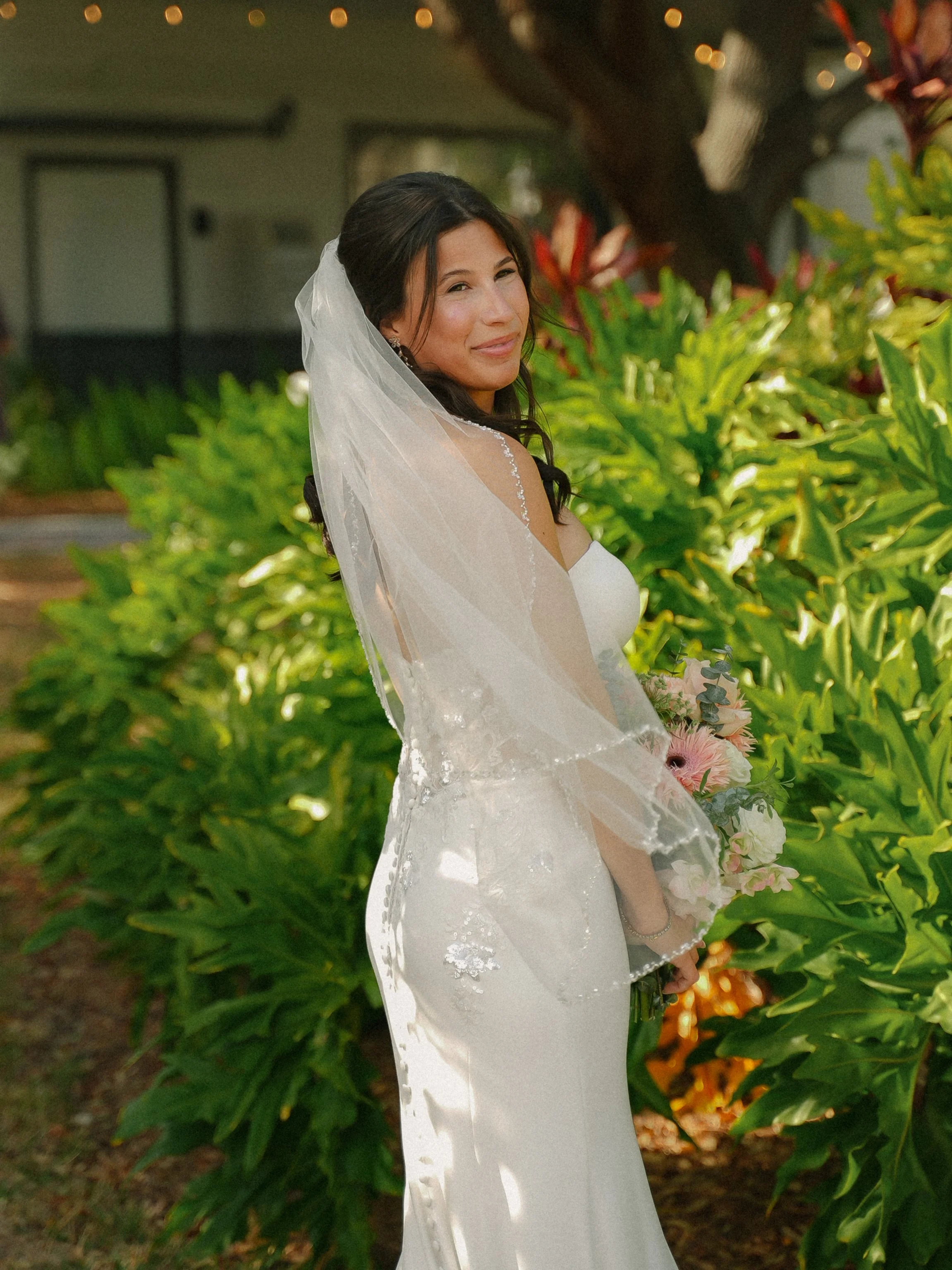 A bride in a white wedding dress with a veil standing outdoors near green plants, holding a bouquet of flowers, smiling at the camera.