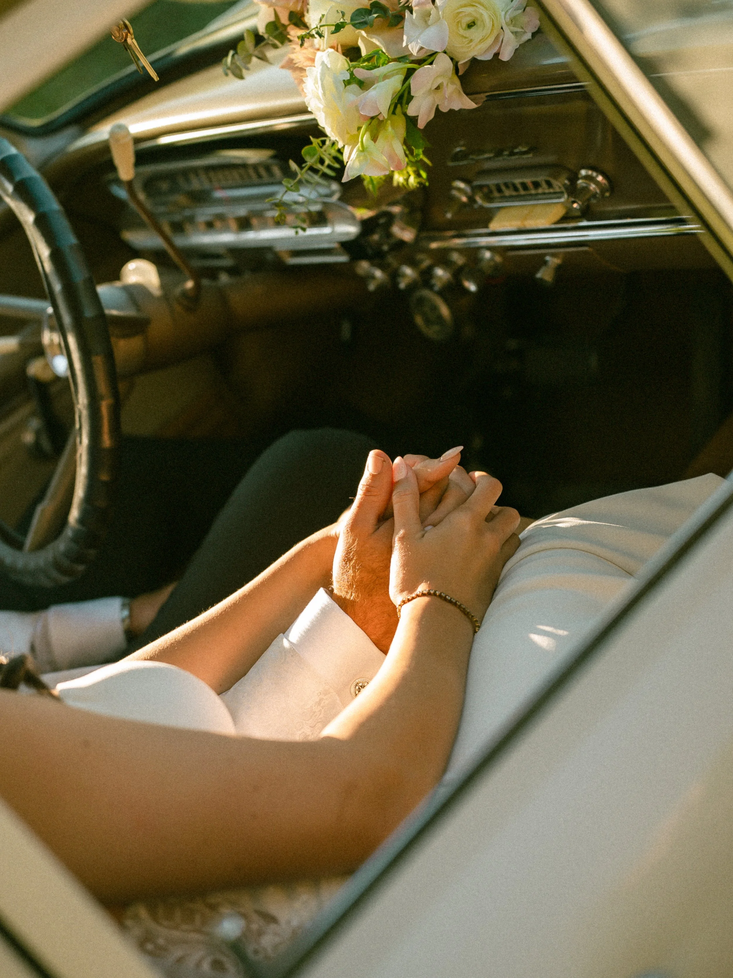A couple holding hands on the front seat of a vintage car decorated with flowers, with the woman wearing a white dress and bracelet, and the man in a white shirt.
