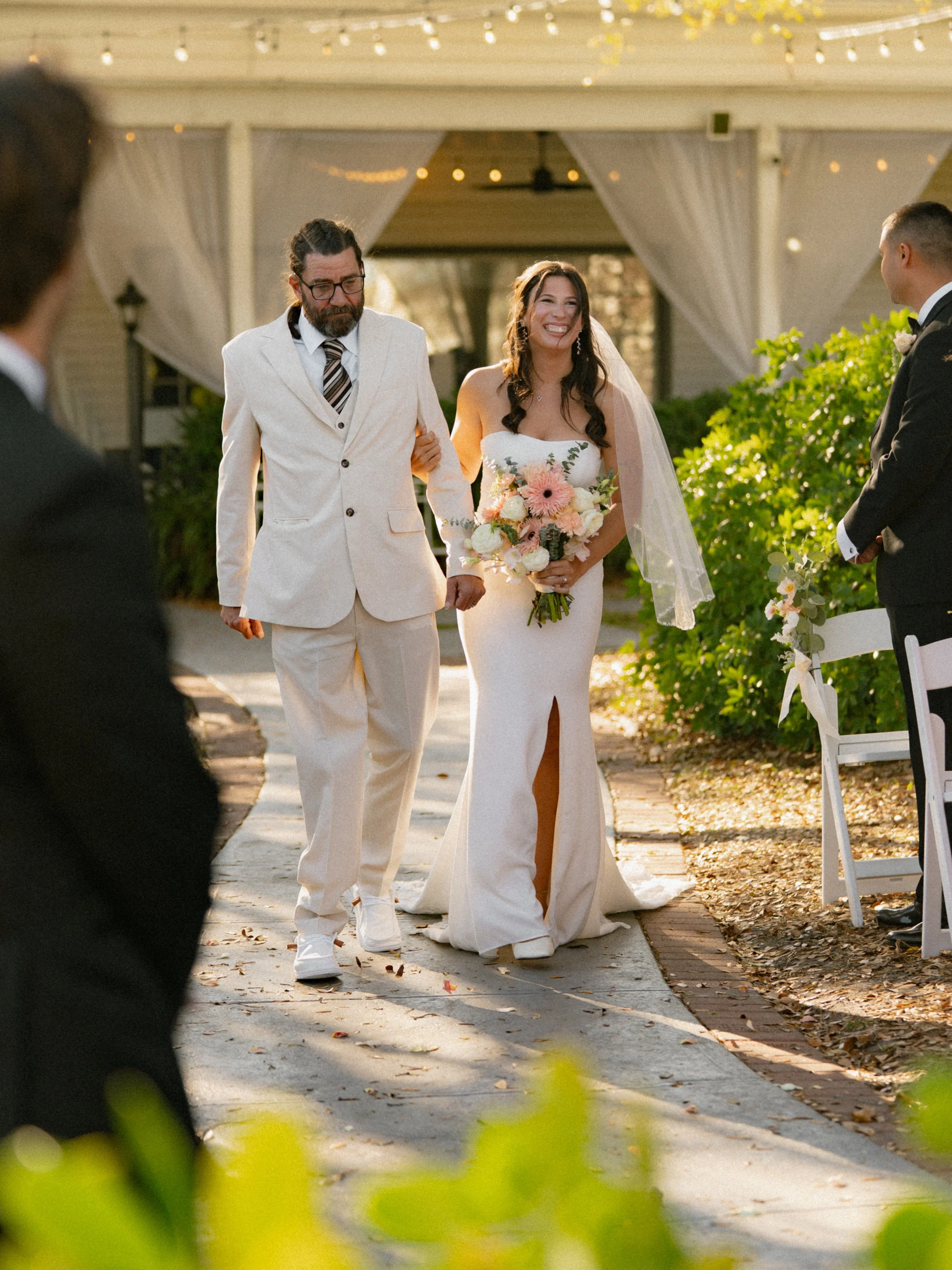 A bride walking down the aisle with her father at an outdoor wedding ceremony, holding a bouquet of pink and white flowers, with groom waiting at the altar.