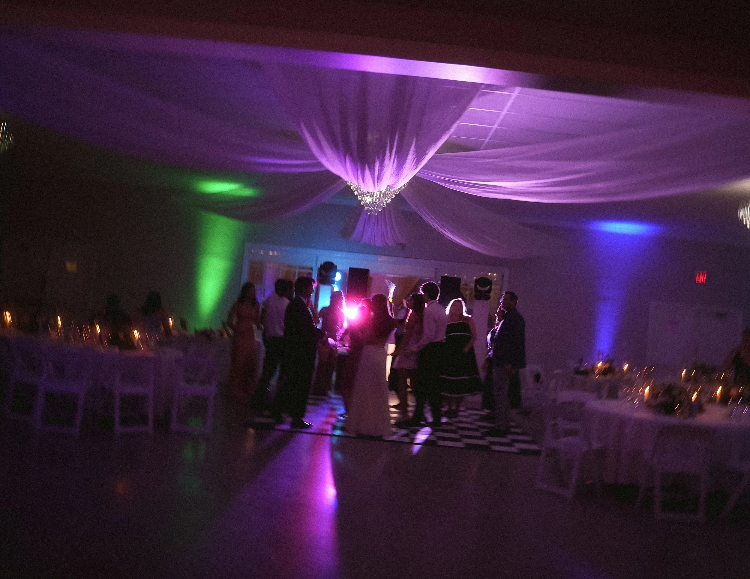 People dancing and socializing at a party or celebration in a dimly lit hall with colorful lighting and a decorated ceiling.