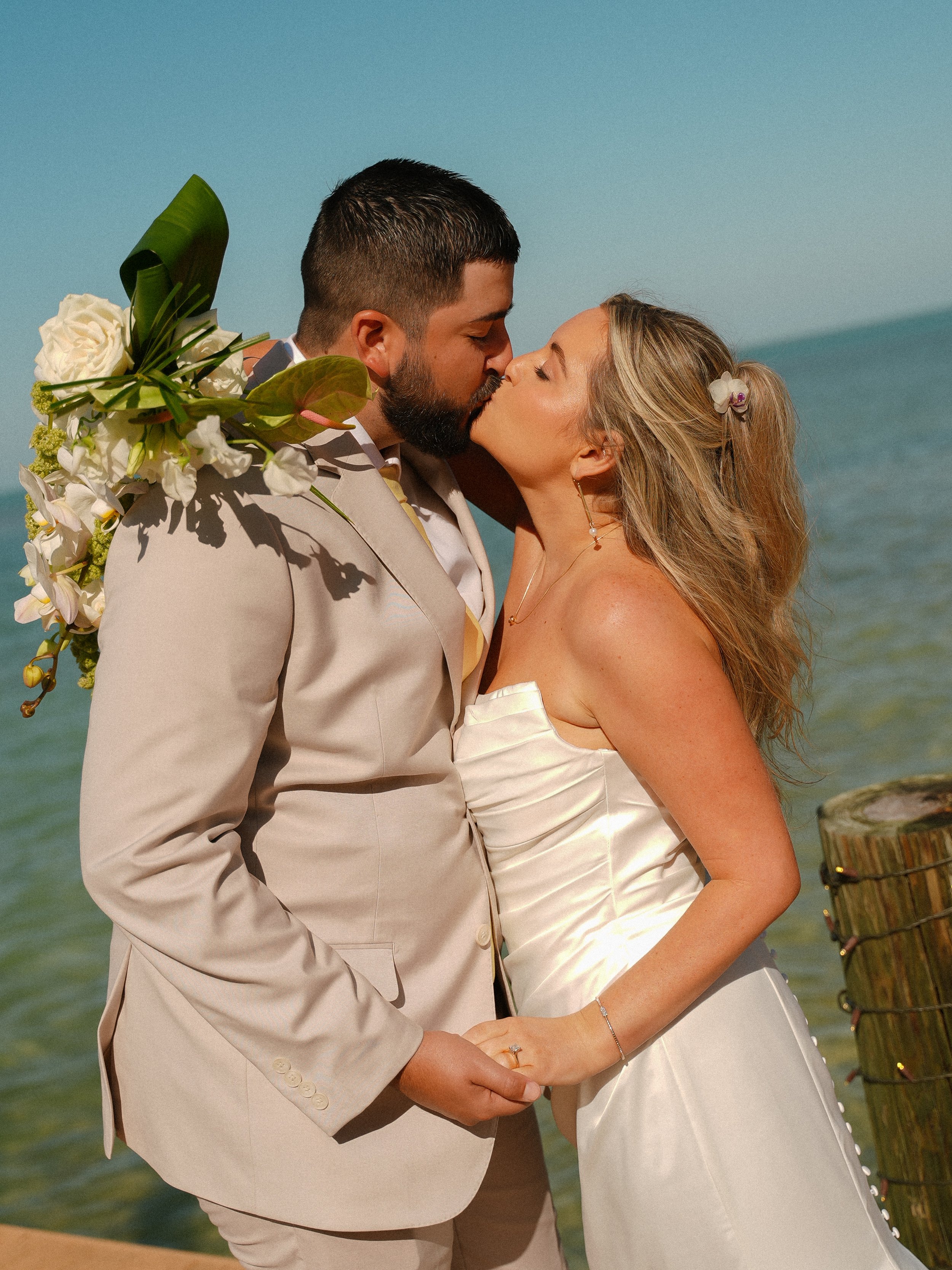 A newlywed couple sharing a kiss at the beach, during their wedding ceremony, with the ocean and sky in the background.