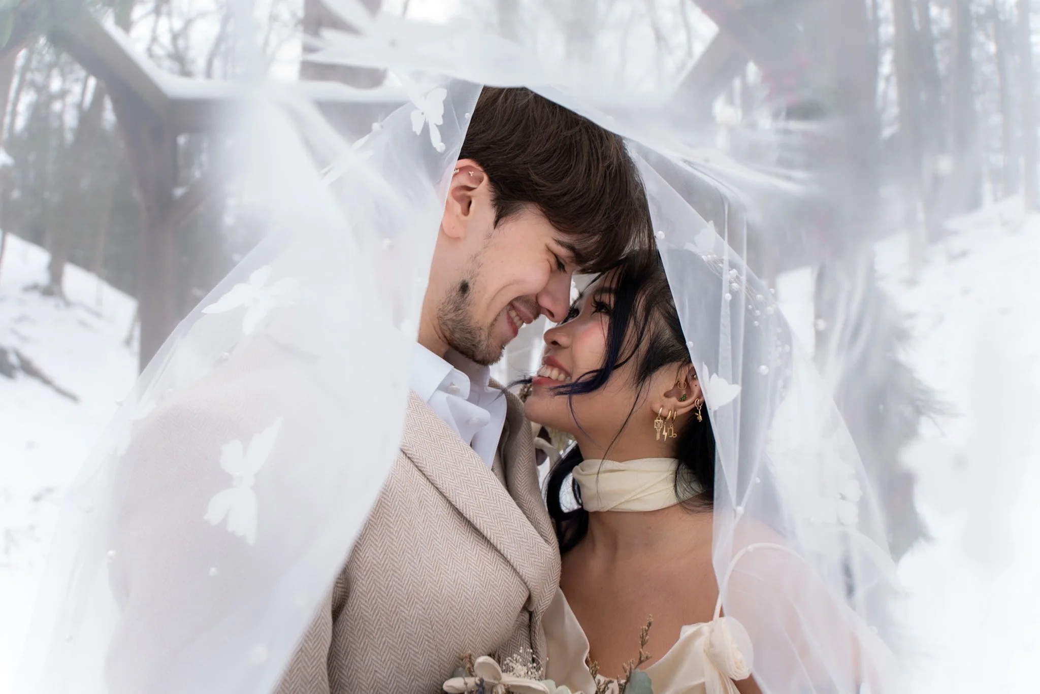 A couple smiling and touching foreheads under a sheer white veil in a snowy outdoor setting.