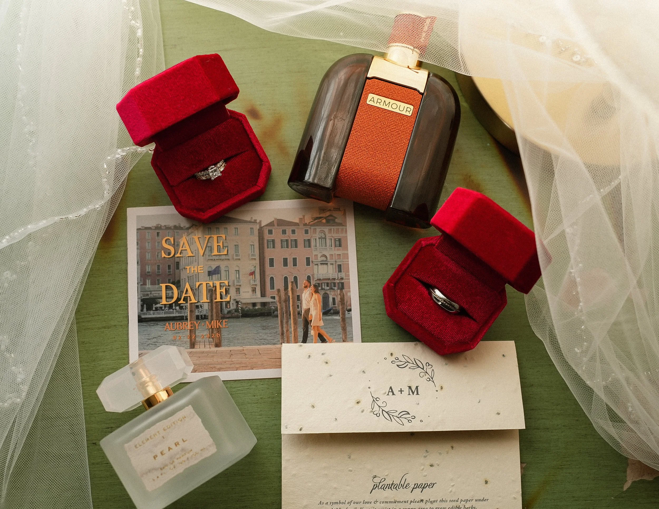 A wedding invitation with the words 'Save the Date,' two red velvet ring boxes with rings, a perfume bottle labeled 'Pearl,' a vintage-style perfume bottle labeled 'Armour,' and a card with initials 'A + M' on a green surface, surrounded by sheer whi