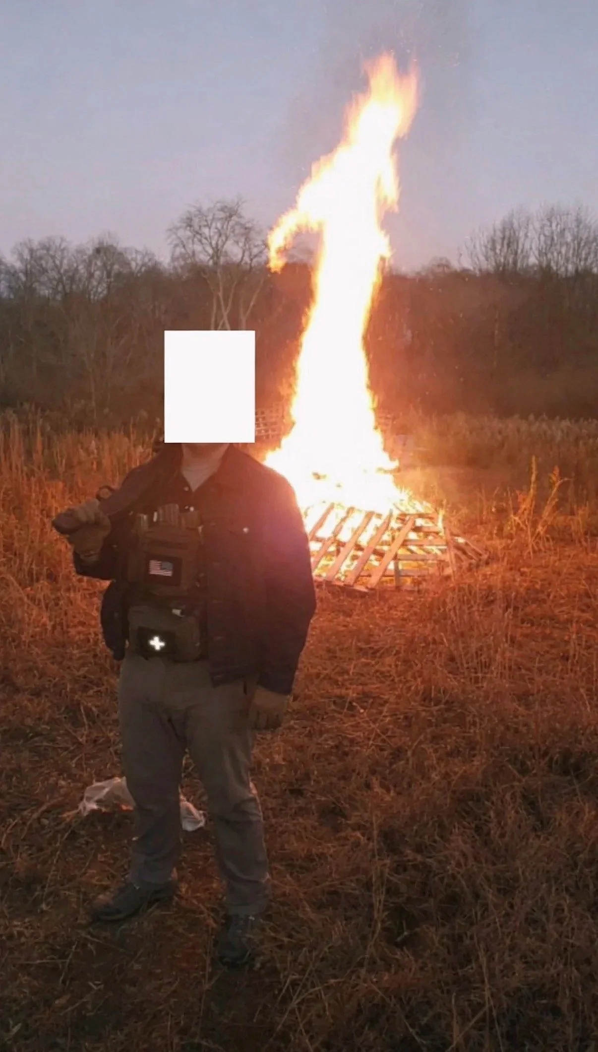 Person standing outdoors at night, in front of a large fire on a grassy field, with trees in the background.