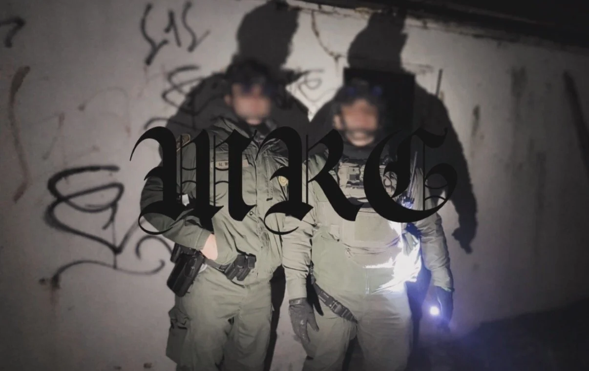 Two people in police or military uniforms standing together in front of a graffiti-covered wall, holding equipment and flashlight.