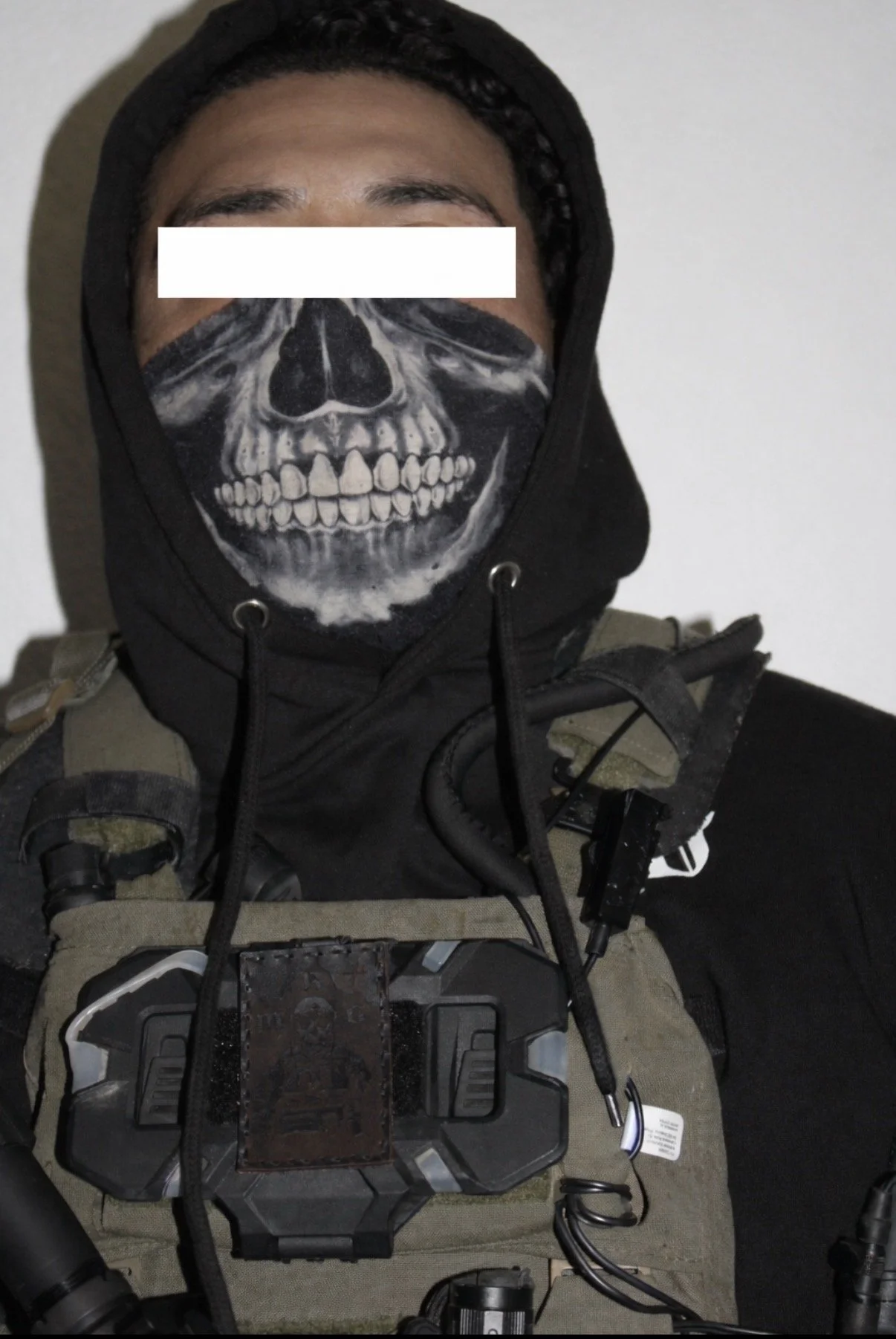 A person wearing a black hoodie and tactical gear, with a skull design face mask covering the lower face.