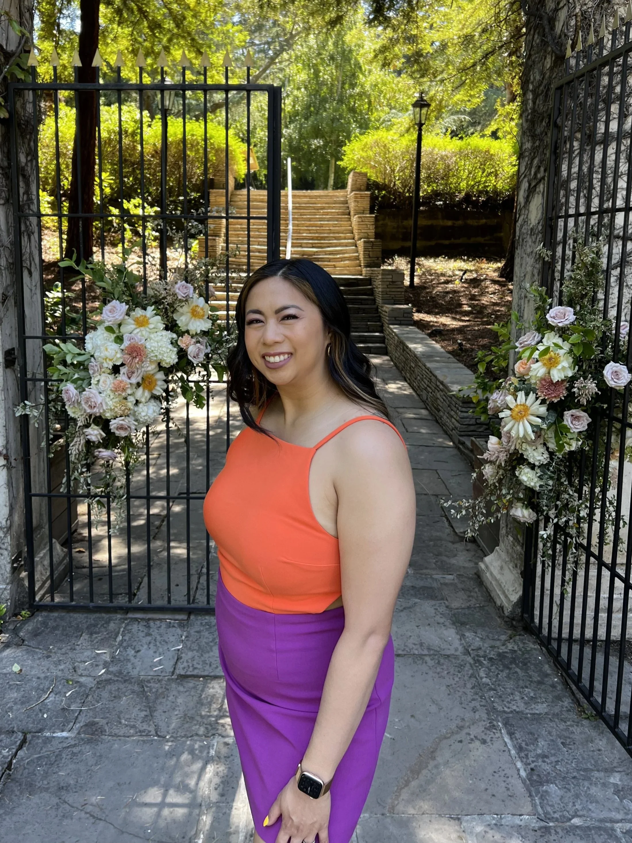 A woman in a sleeveless orange top and purple skirt standing in front of a decorative gate with flowers on either side. She is smiling and wearing a smart watch, with a garden and steps in the background.