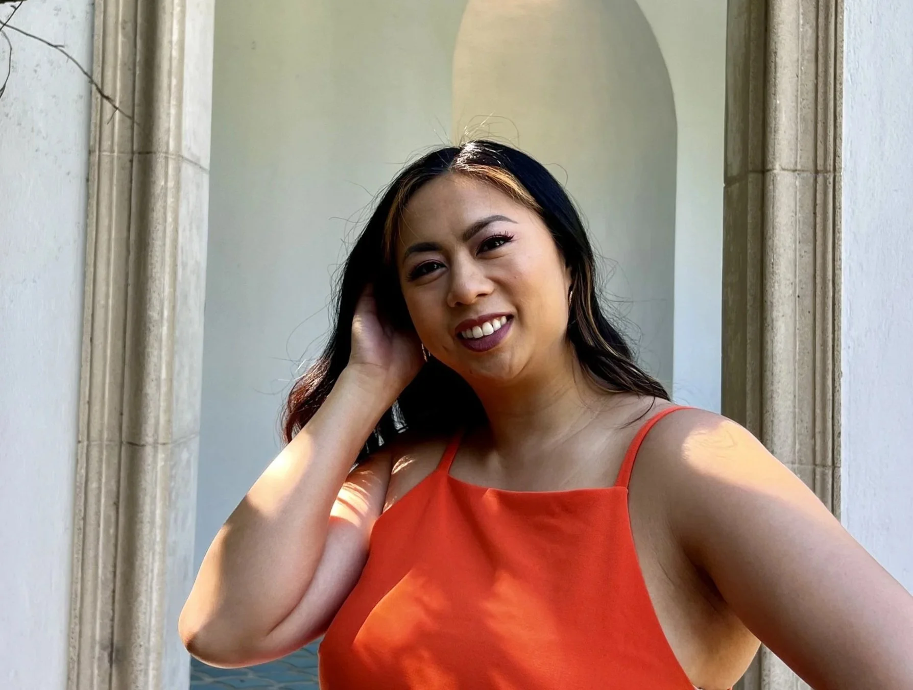 A woman with long black hair and tan highlights wearing an orange spaghetti strap dress, smiling and touching her hair.