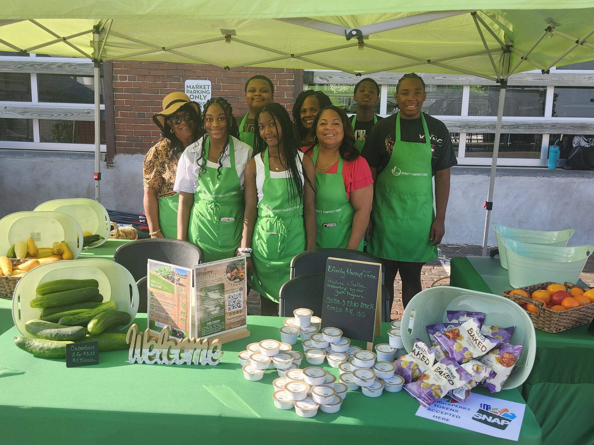 Efinity Harvest Farm team at an outdoor farmers market in Ohio wearing branded aprons, displaying fresh cucumbers, value added products, and local goods under a canopy to support community food access