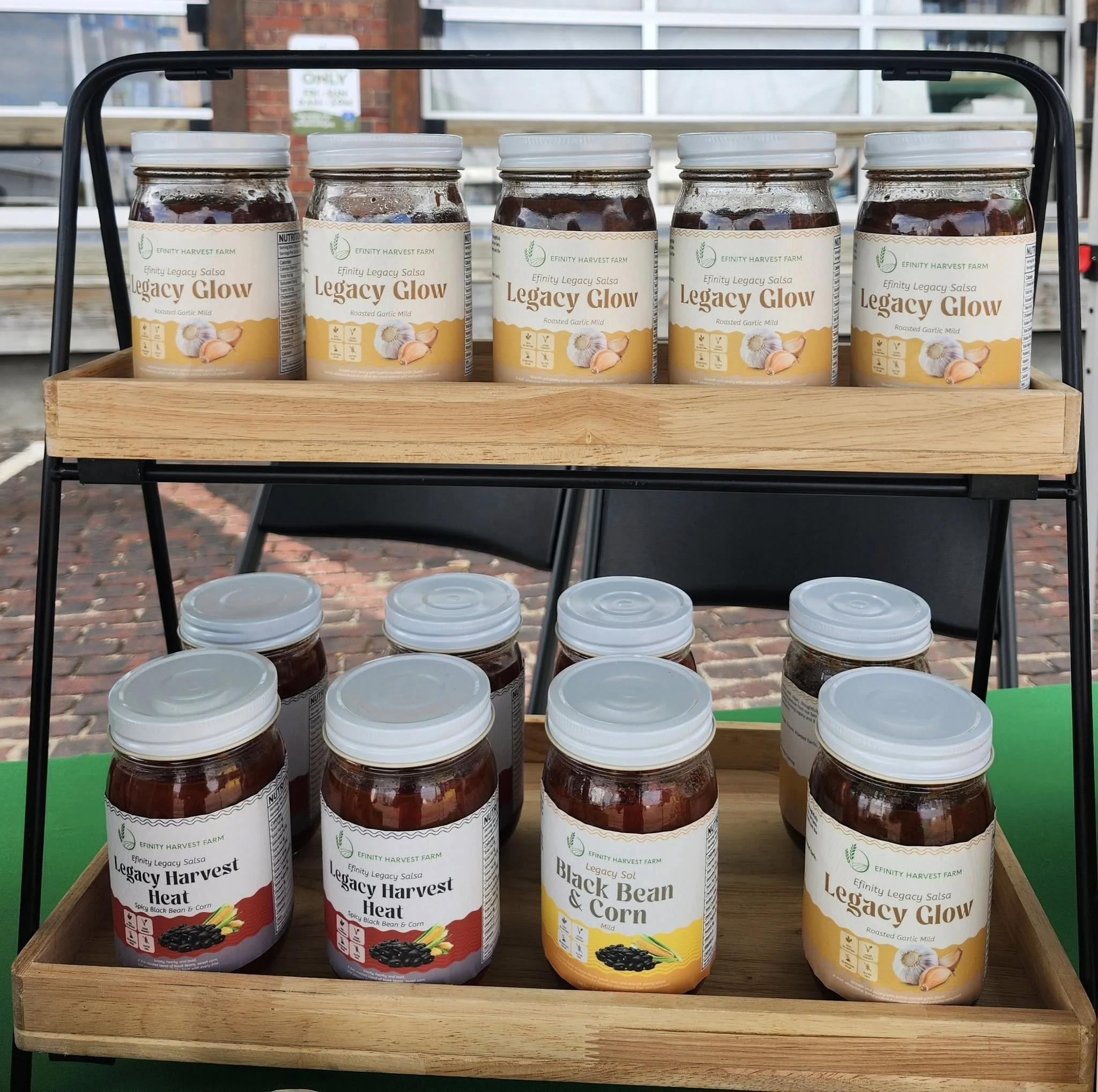 Efinity Harvest Farm Legacy Glow roasted garlic salsa and Legacy Harvest Heat black bean and corn salsa displayed on wooden shelves at a farmers market in Trotwood, Ohio featuring locally made value added products