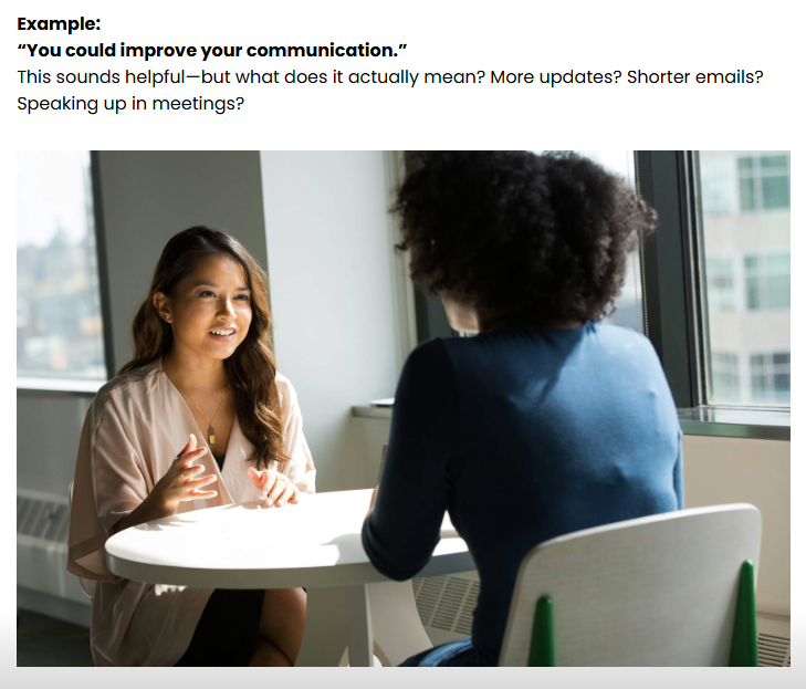 Two women having a conversation at a white table near a window in a modern office setting, with city buildings visible outside.
