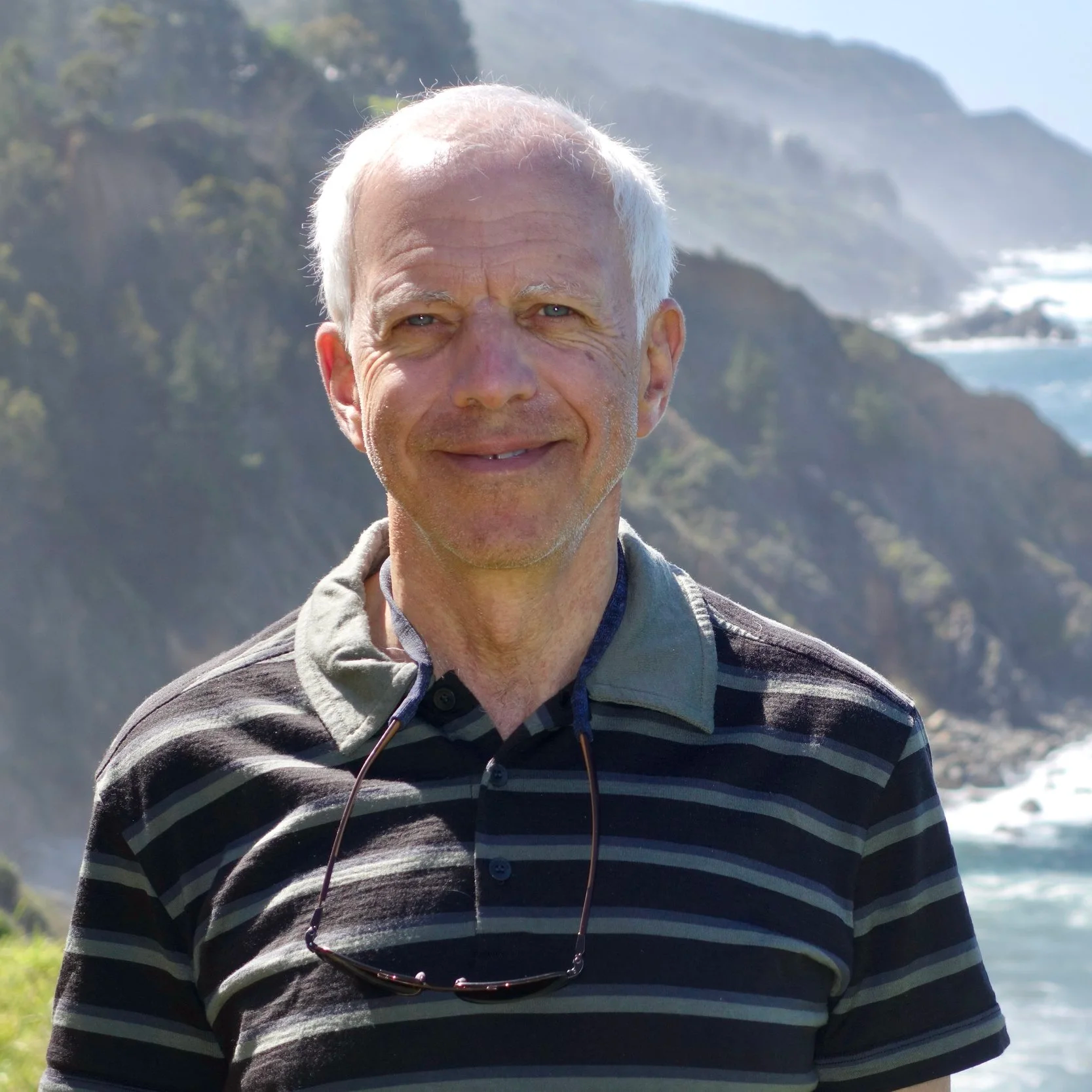 An older man with short gray hair smiling outdoors with a scenic coastal background that includes cliffs, greenery, and ocean waves.