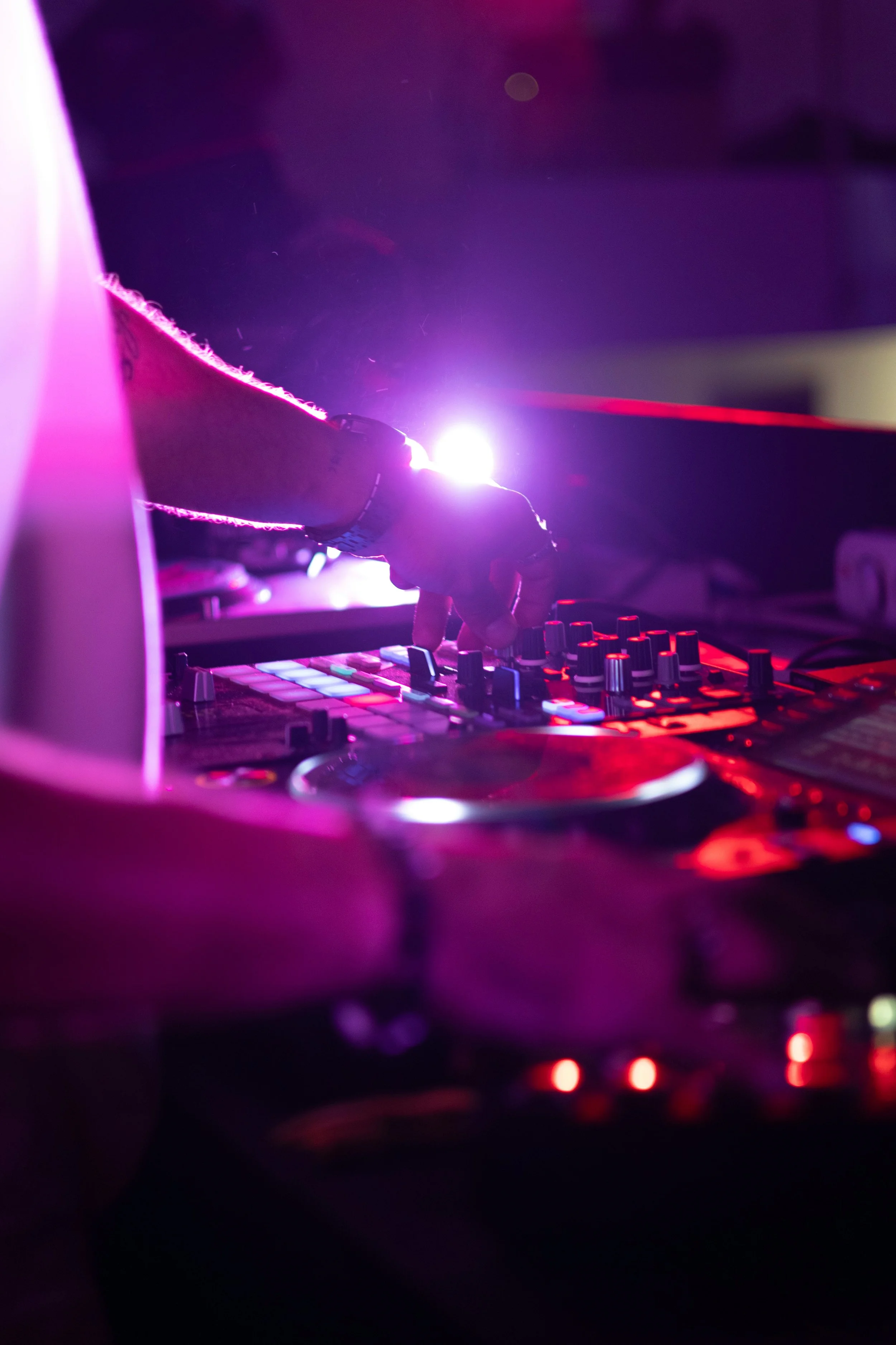 Close-up of a DJ's hand adjusting controls on a mixing console illuminated by vibrant pink and purple lights, with a bright light source in the background creating a glow.