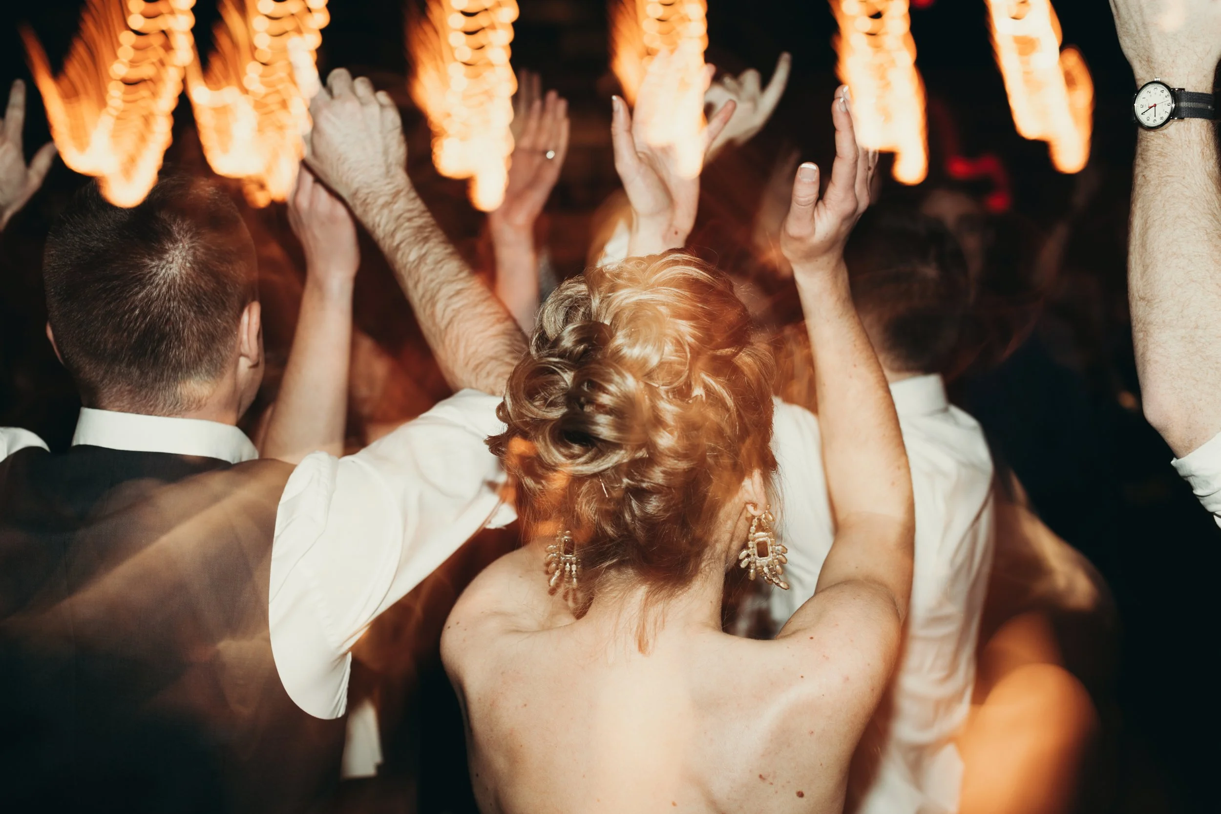 A group of people dancing closely together at a party or celebration, with their hands raised in the air, lit by warm, orange lighting.