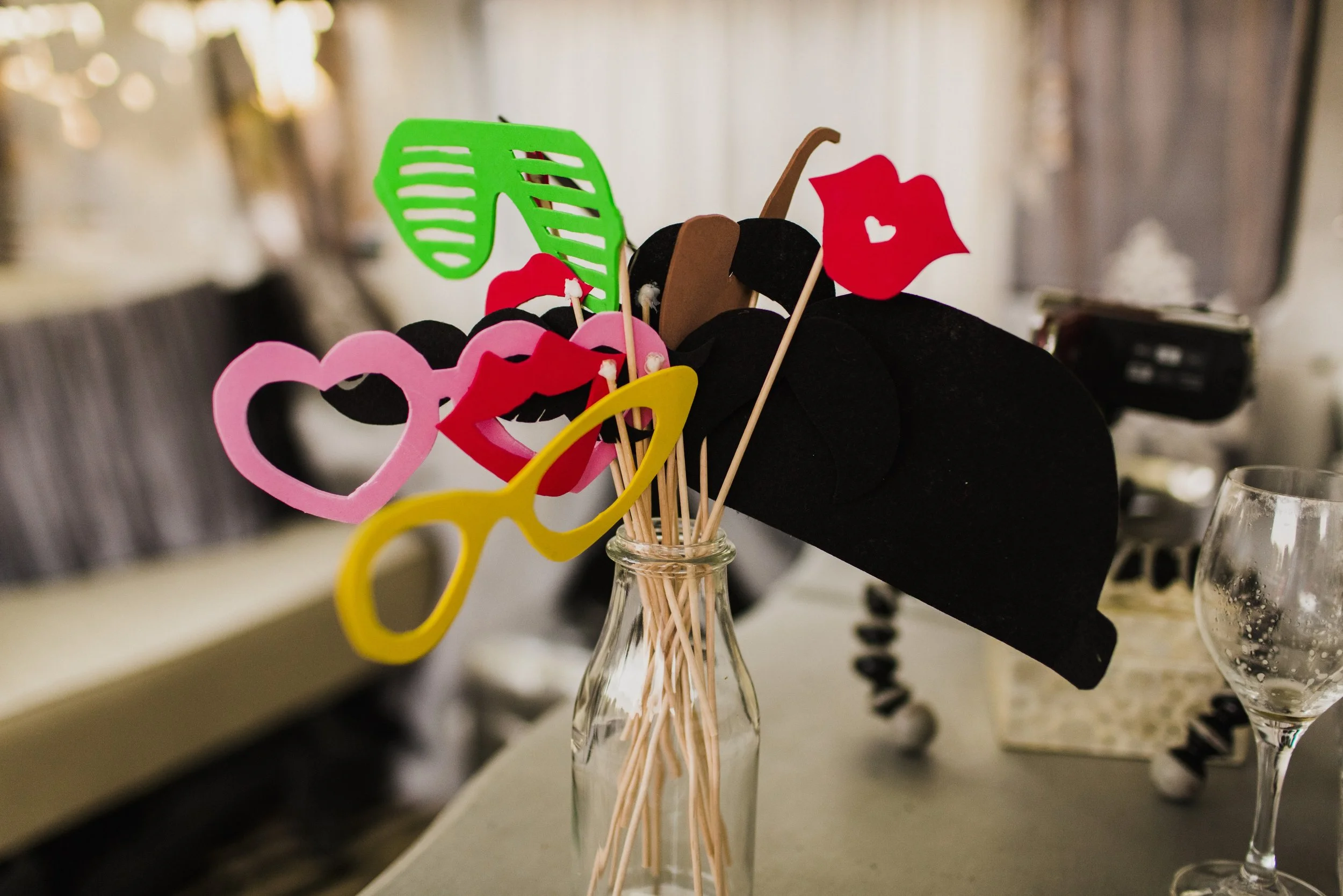 Colorful novelty glasses and props in a glass jar on a table, possibly for a celebration or photo booth, with blurred background.