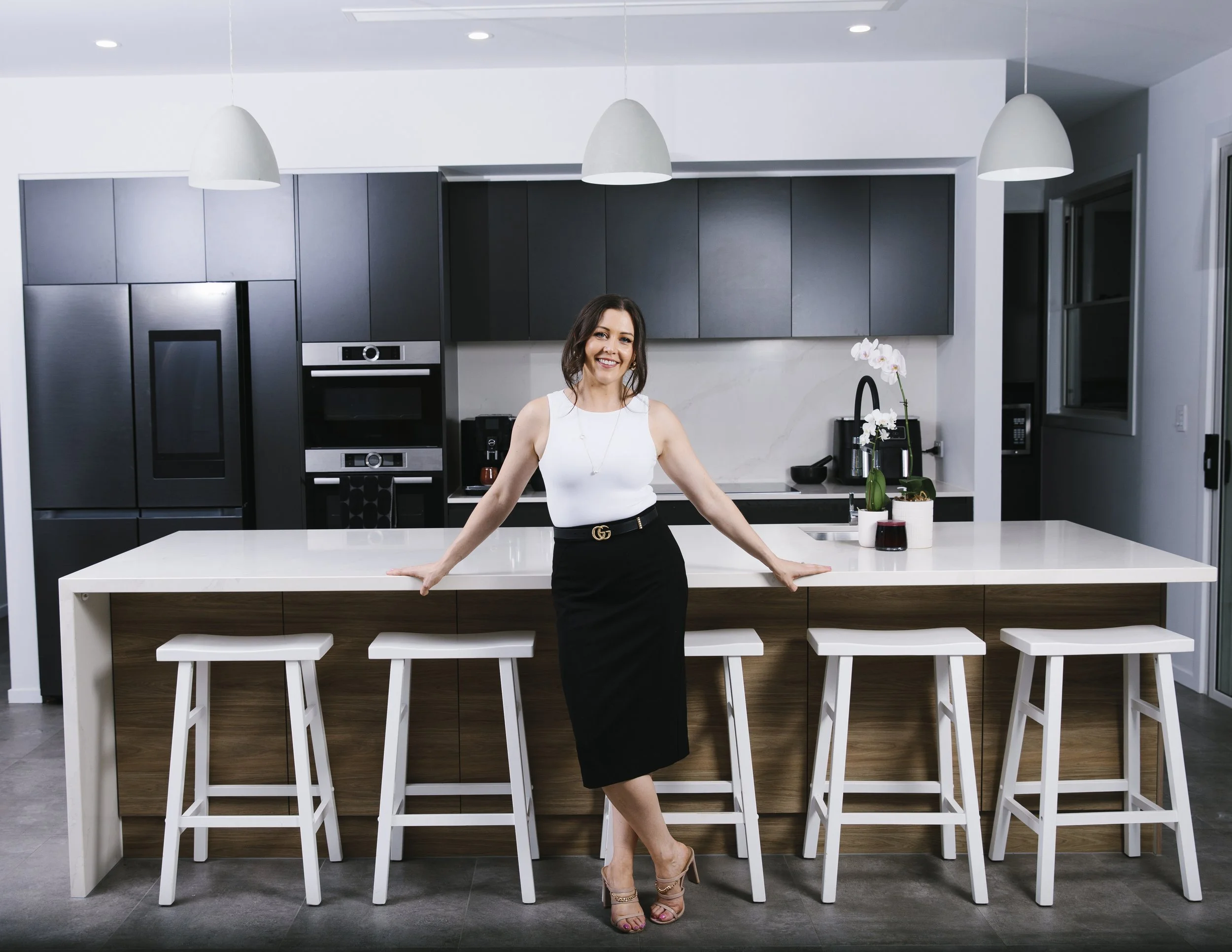 Woman standing at a modern kitchen island, smiling, with black and white cabinetry, three pendant lights, and decorative plants on the counter.