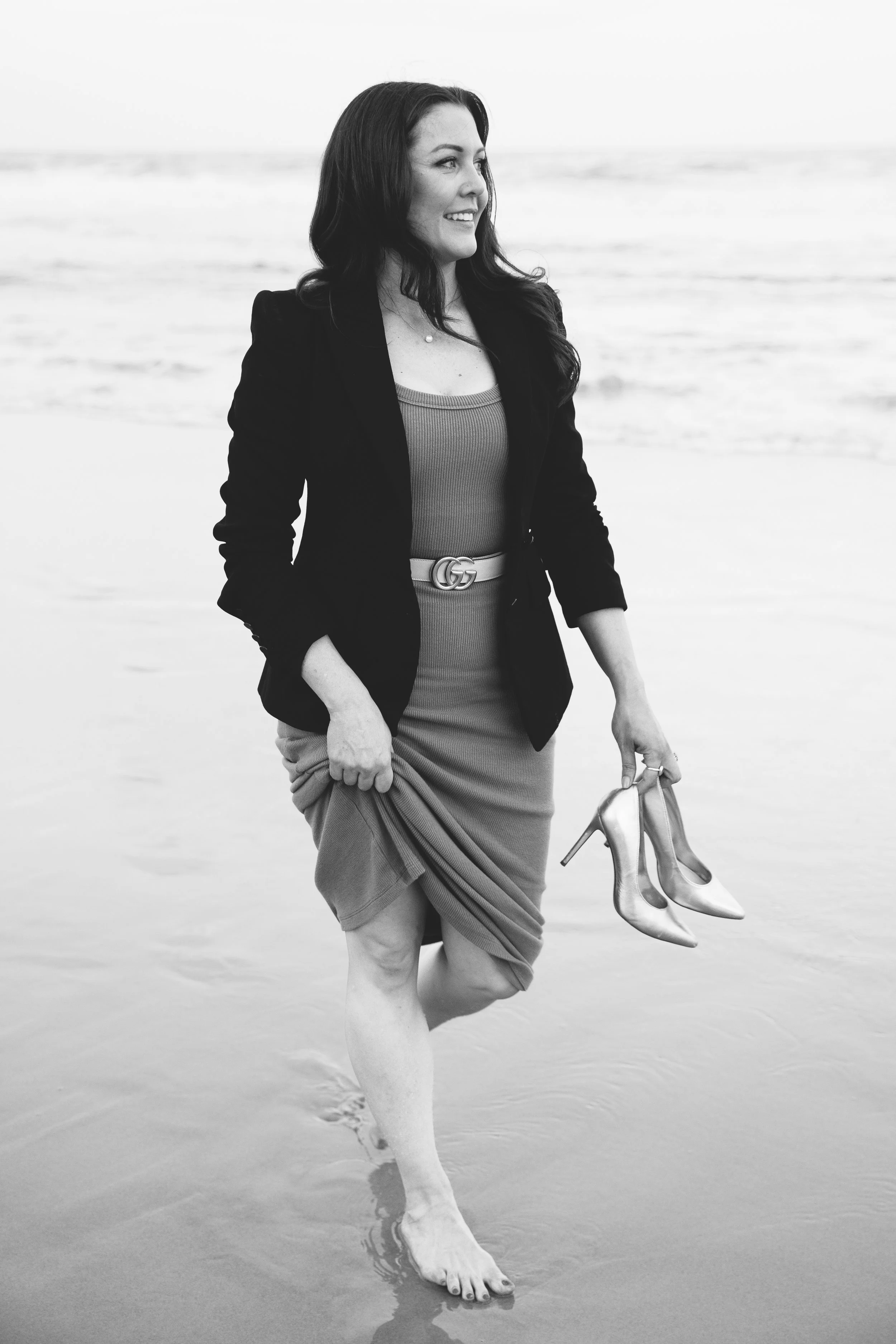 A woman walking barefoot on the beach, holding a pair of high-heeled shoes, wearing a blazer and a dress, smiling and looking to the side.