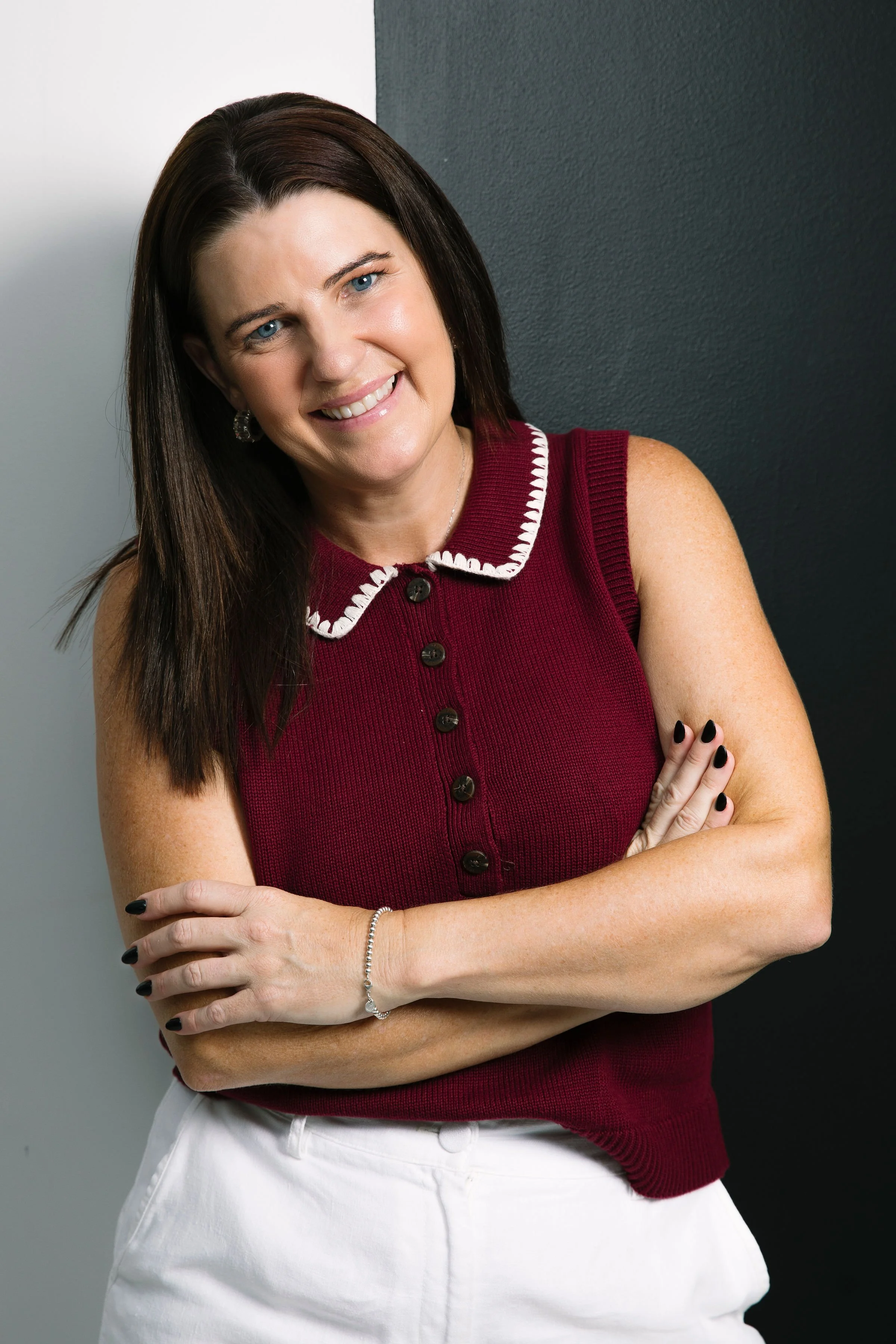 Woman with dark brown hair, wearing a sleeveless maroon sweater with black buttons and white embroidered collar, smiling and crossing arms, standing against a dark wall.