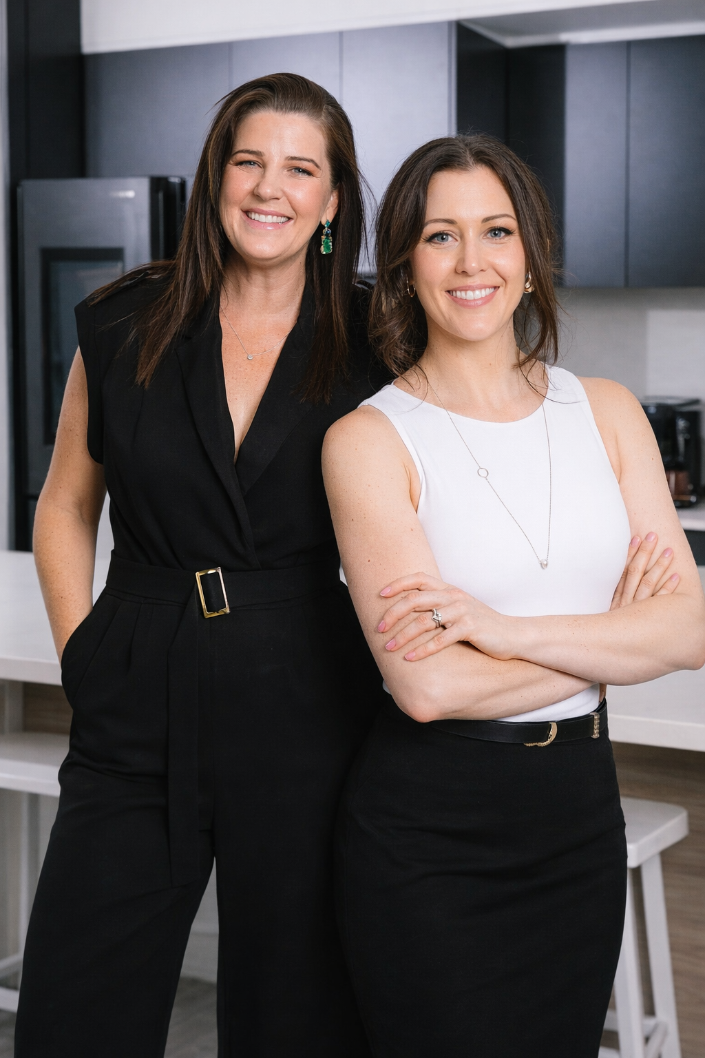 Two women standing in a modern kitchen, smiling, one in a black sleeveless jumpsuit with a belt, the other in a white sleeveless top and black skirt with her arms crossed.