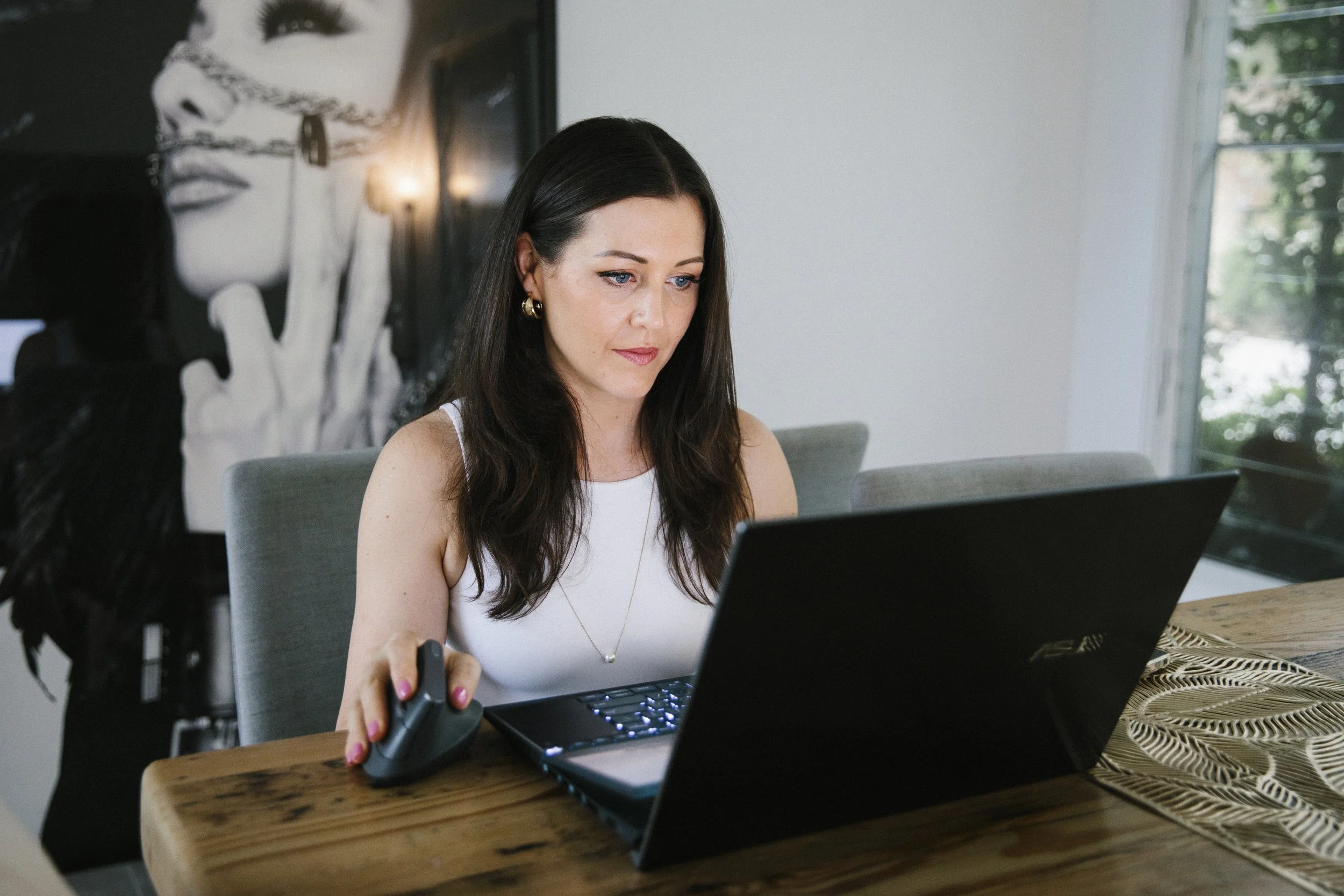 A woman with long dark hair and earrings is sitting at a wooden table, working on a laptop with a wired mouse. She is in a bright room with a large window, and a black-and-white portrait of a woman with finger on lips drawing on the wall behind her.