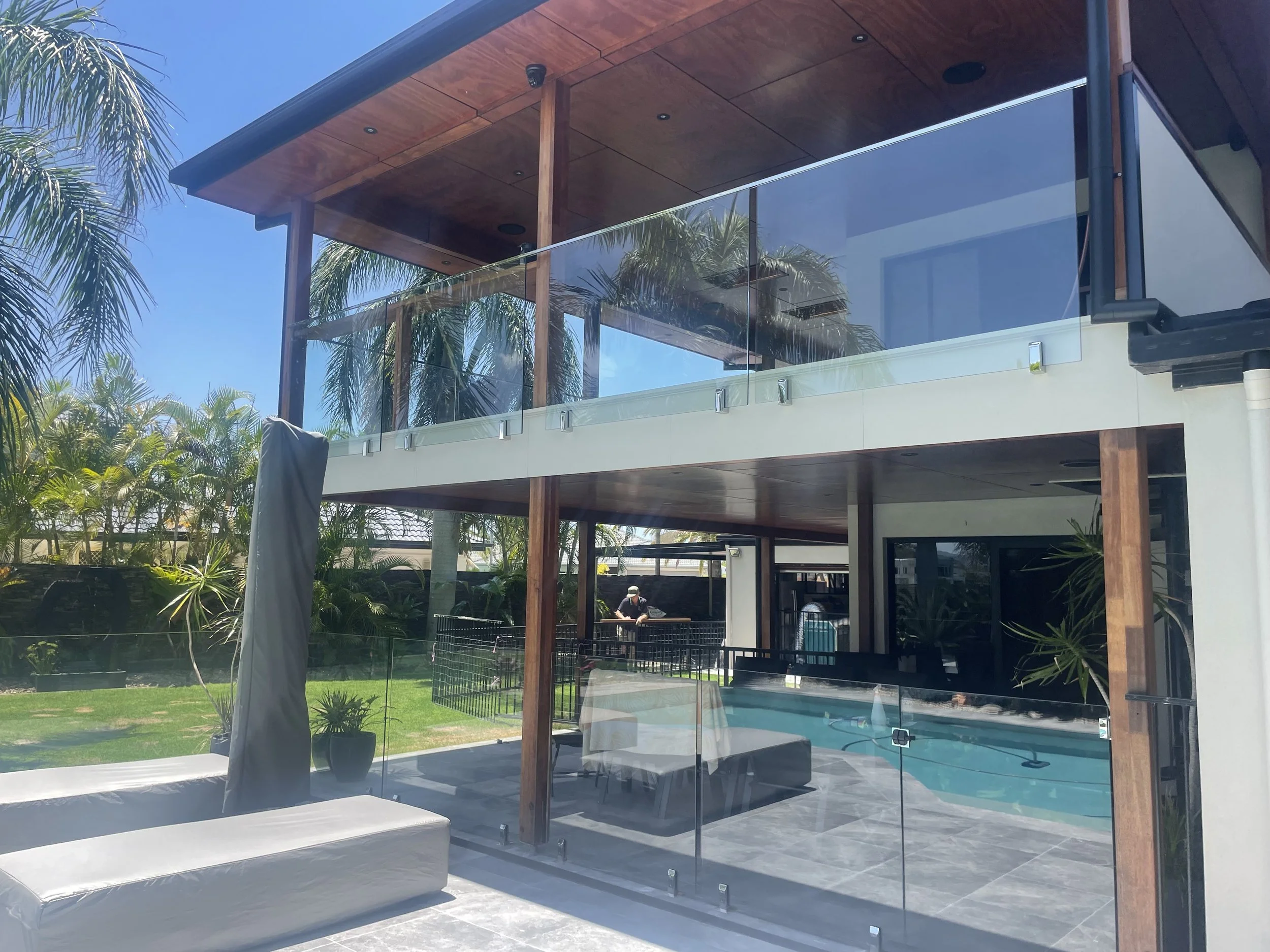 Modern house with a glass balcony, wooden roof, and outdoor area with greenery and palm trees.