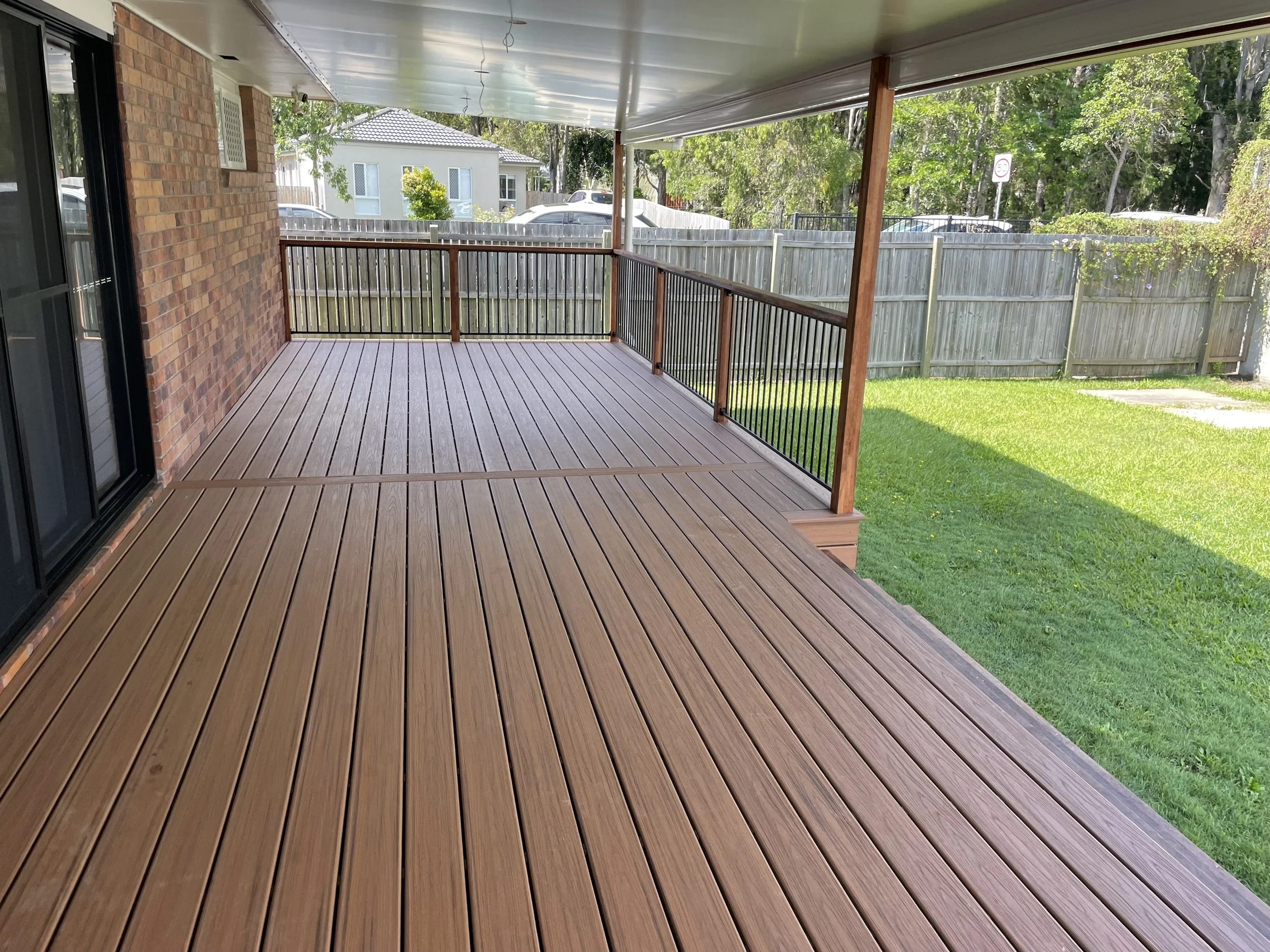 A covered porch with wooden decking, black railing, brick exterior wall, and view of a backyard with green grass, a wooden fence, trees, and neighboring houses.