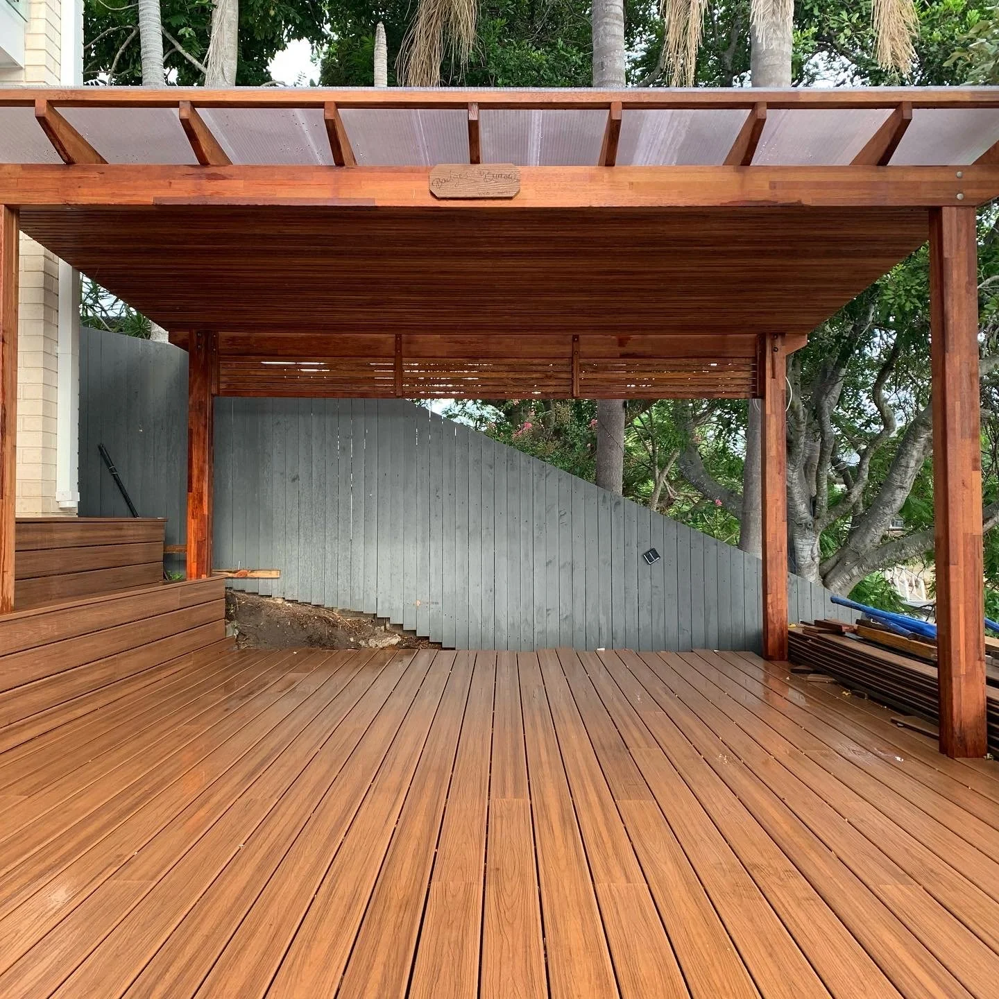 Newly built wooden deck with a shaded upper level, surrounded by green trees and a gray fence in the background.