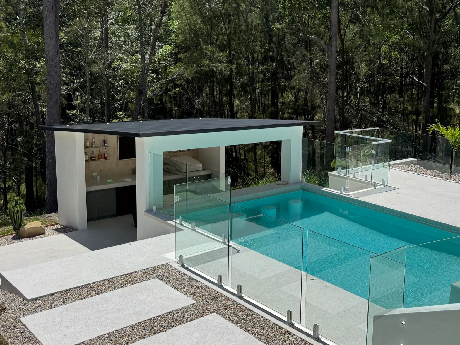 Modern backyard with a swimming pool, glass fencing, outdoor shower, and small pool house with a bar area, surrounded by trees.