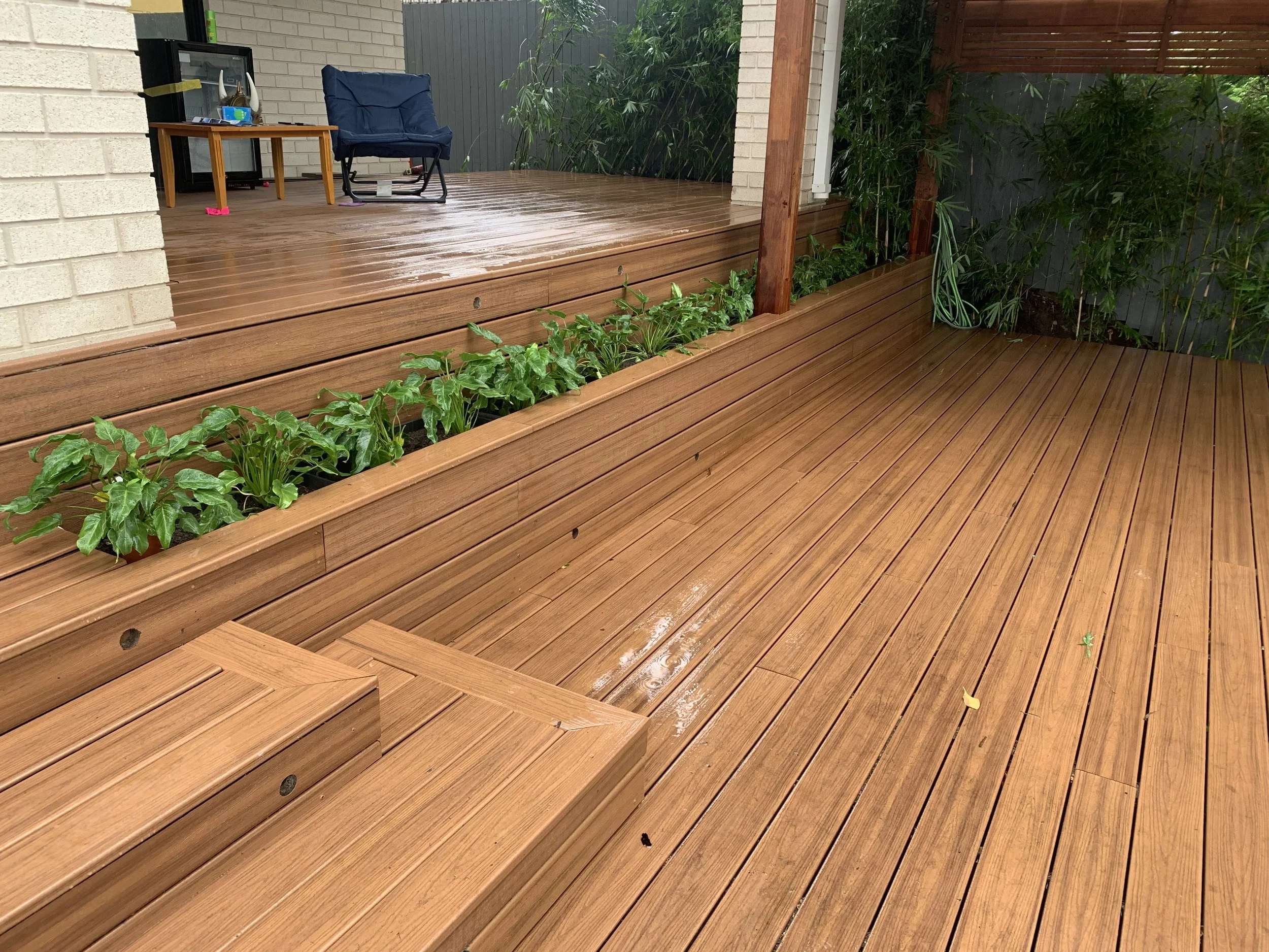 A wet wooden deck with stairs, a blue chair, a small wooden table, some green plants, a brick wall on the left, and a wooden fence and foliage in the background.
