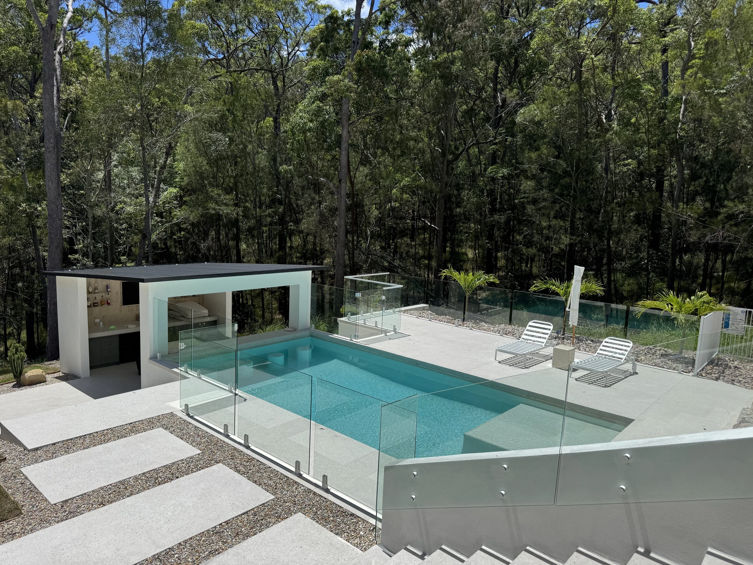 Modern backyard with a swimming pool enclosed by glass fencing, surrounded by a view of tall trees and lush greenery, with a shaded outdoor bar area and lounge chairs.