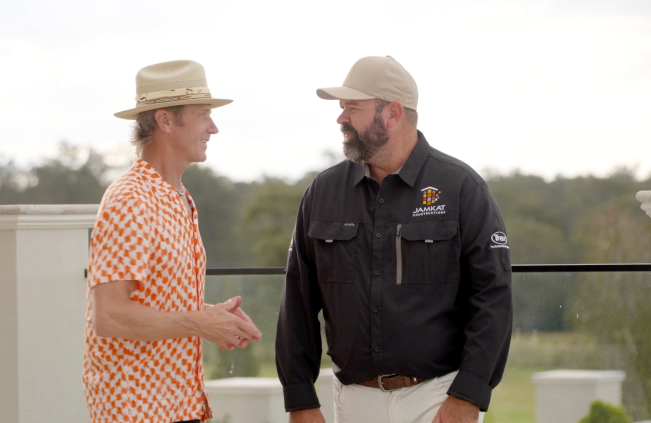 Two men, one in a beige hat and patterned orange shirt, and the other in a beige cap and black shirt with a logo, are smiling and looking at each other outdoors.