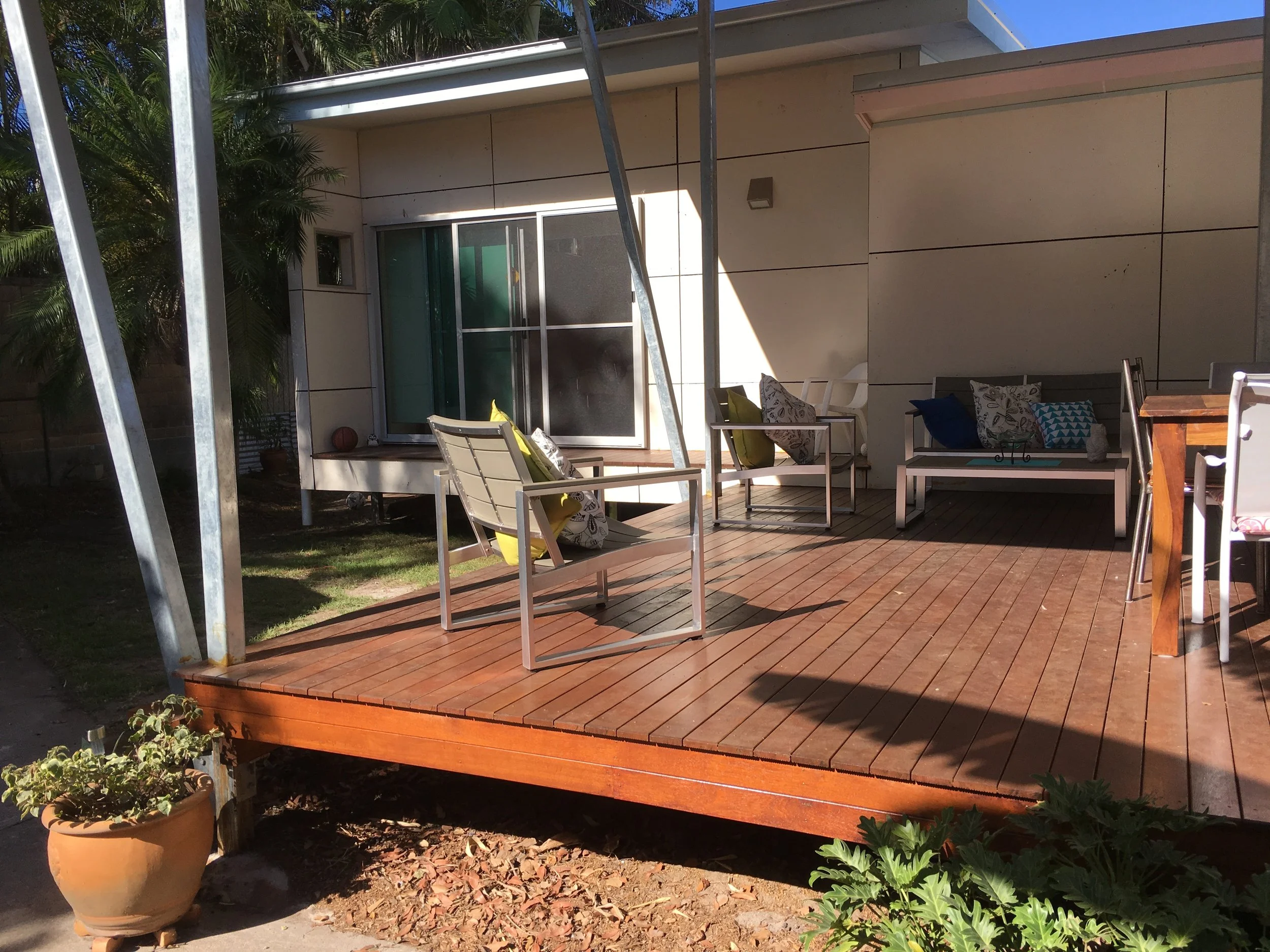 View of a backyard patio with a wooden deck, metal patio furniture with cushions, a potted plant, and a sliding glass door leading into a house. There are trees and plants surrounding the area.