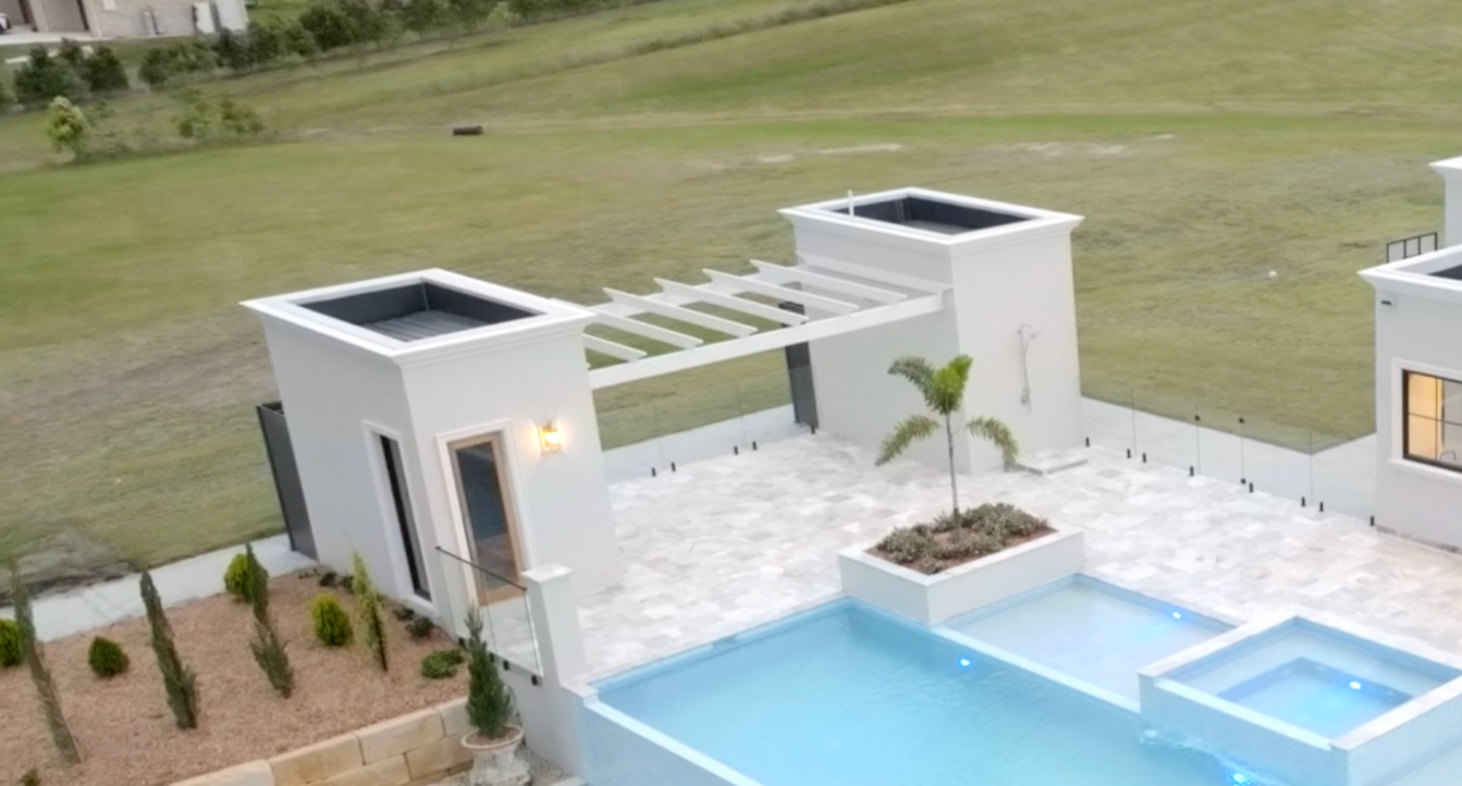 Modern backyard with a swimming pool, hot tub, small tree, and outdoor patio area with white buildings and a grassy yard in the background.