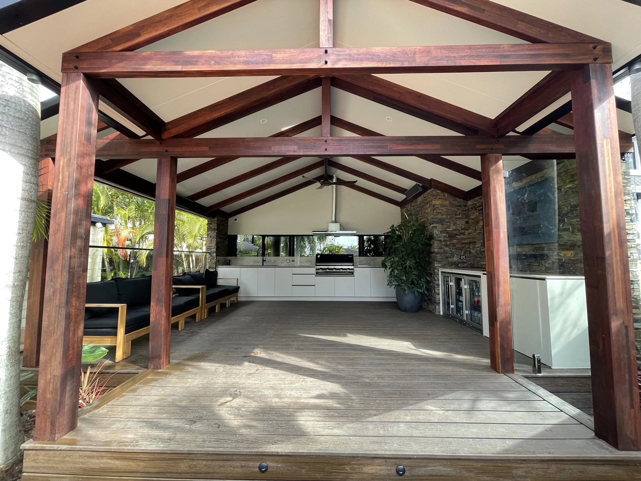 Open outdoor pavilion with a wooden frame, ceiling fan, and kitchen area visible inside, surrounded by tropical plants.