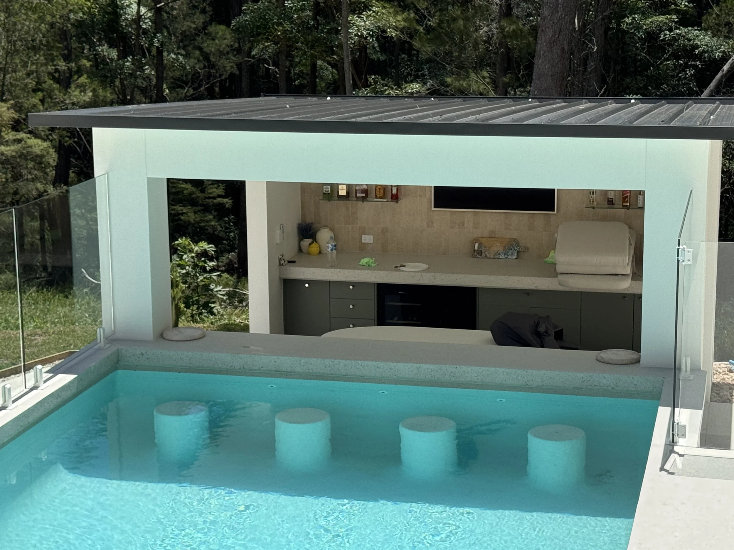 Modern outdoor pool with turquoise water, surrounded by glass fencing, view of a house interior with a kitchen area, and lush green trees in the background.