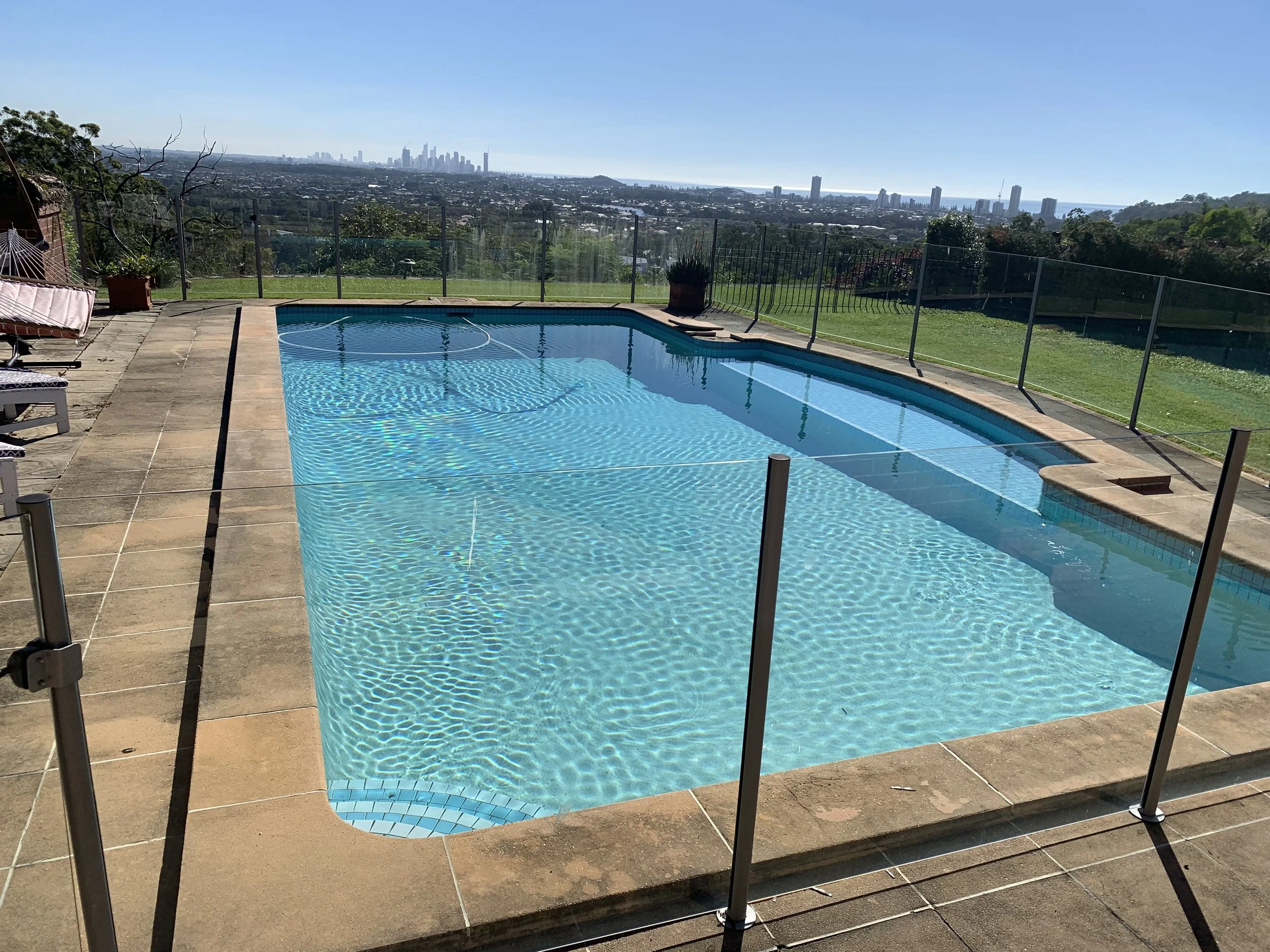 Swimming pool with a city skyline in the background, enclosed by a glass fence, on a sunny day.