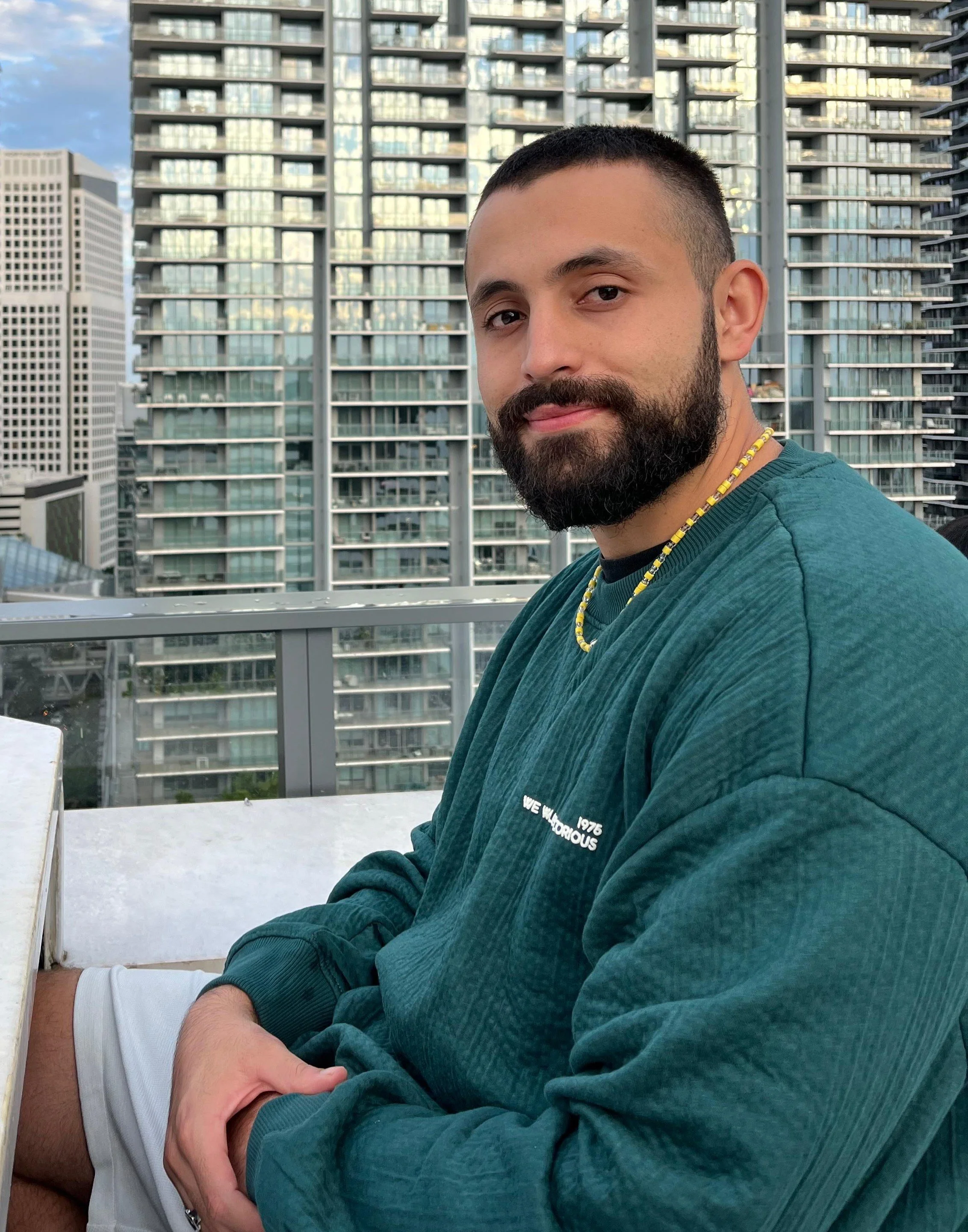 Hombre con barba y camiseta azul sentado en un balcón con vista a edificios modernos en una ciudad.