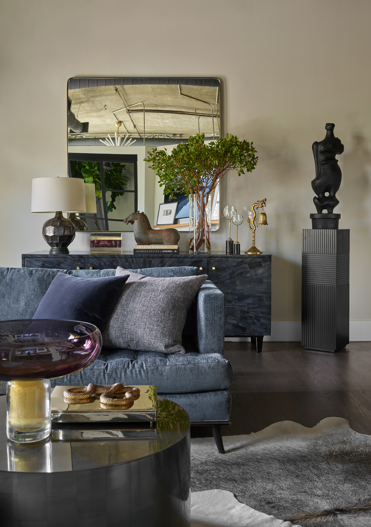 Living room with a dark gray sofa, a black and gold coffee table, a gold and glass tray with a woven object, a lamp with a textured dark base, a large mirror, a black sideboard with a plant, art pieces, and decorative objects, a tall black sculpture, and a gray textured rug.