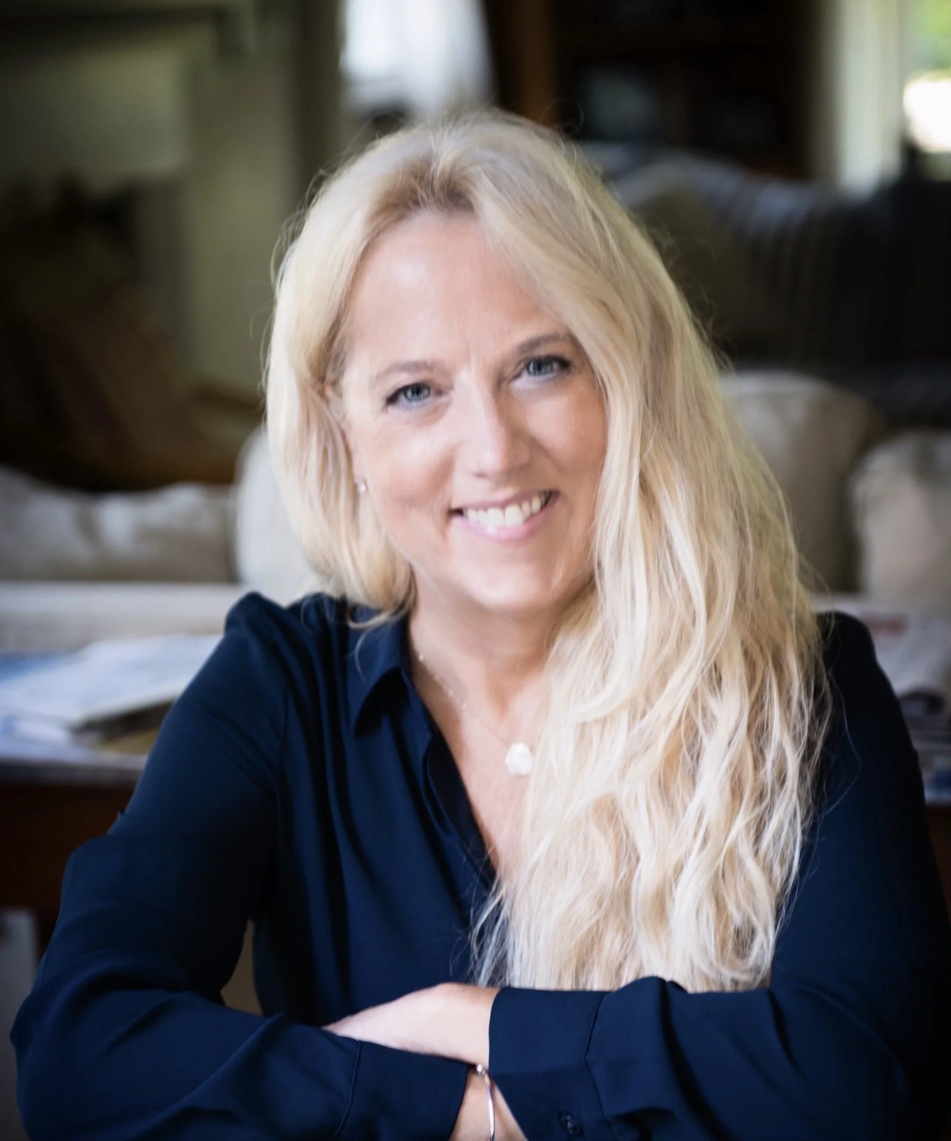 A woman with long blonde hair, blue eyes, and a big smile, wearing a dark blue top, is sitting indoors with her arms crossed on a table, with a blurred background of a living room with a couch and window.